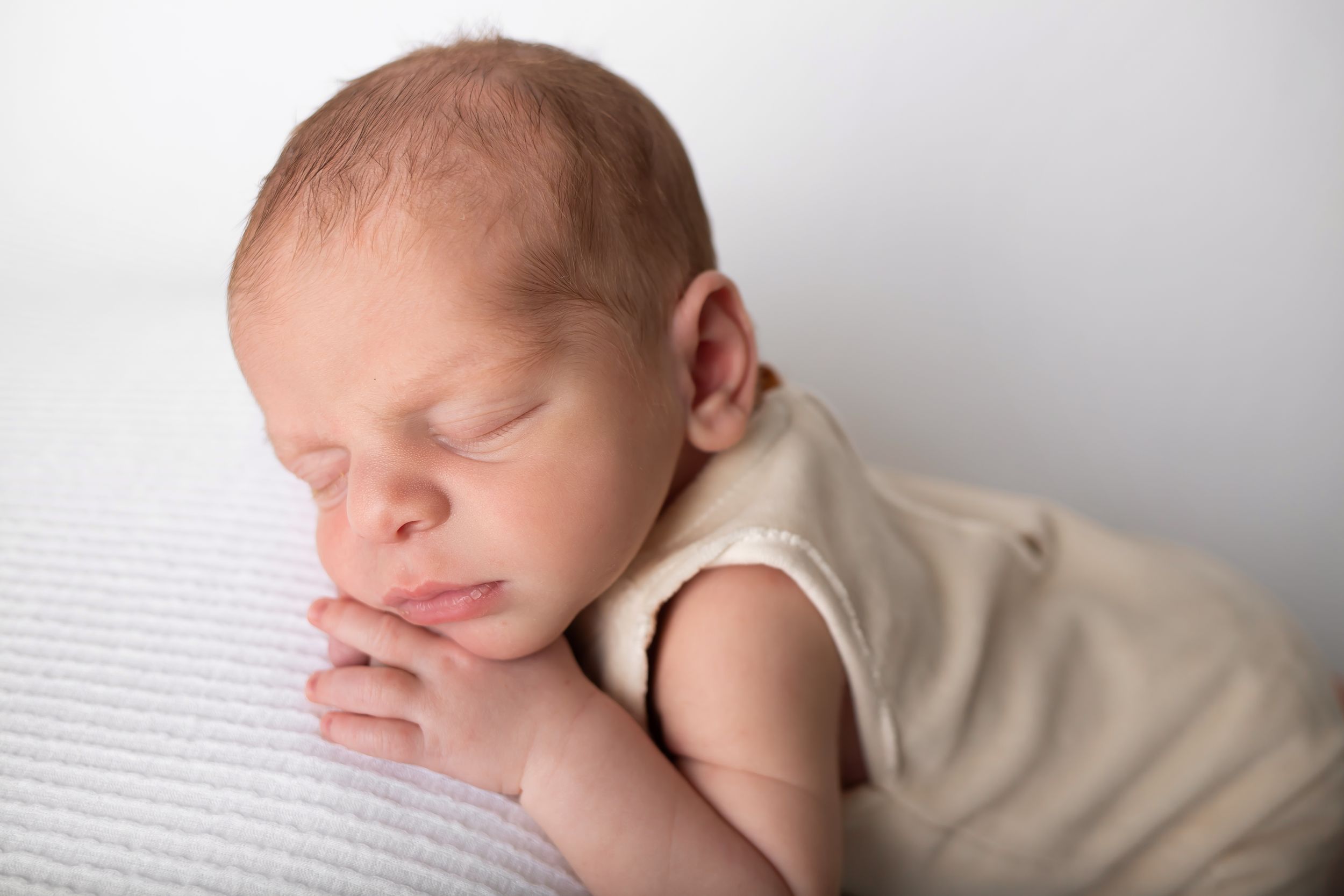 Newborn baby sleeping with hands folded under chin on textured white surface.