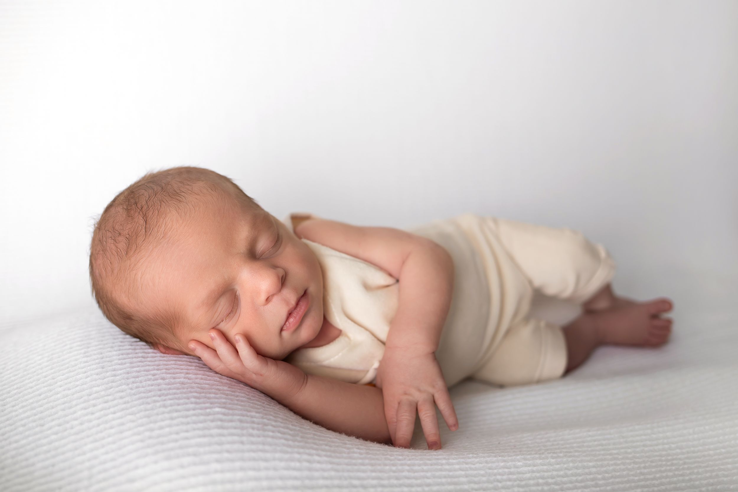 Sleeping newborn in cream colored outfit resting on white background.