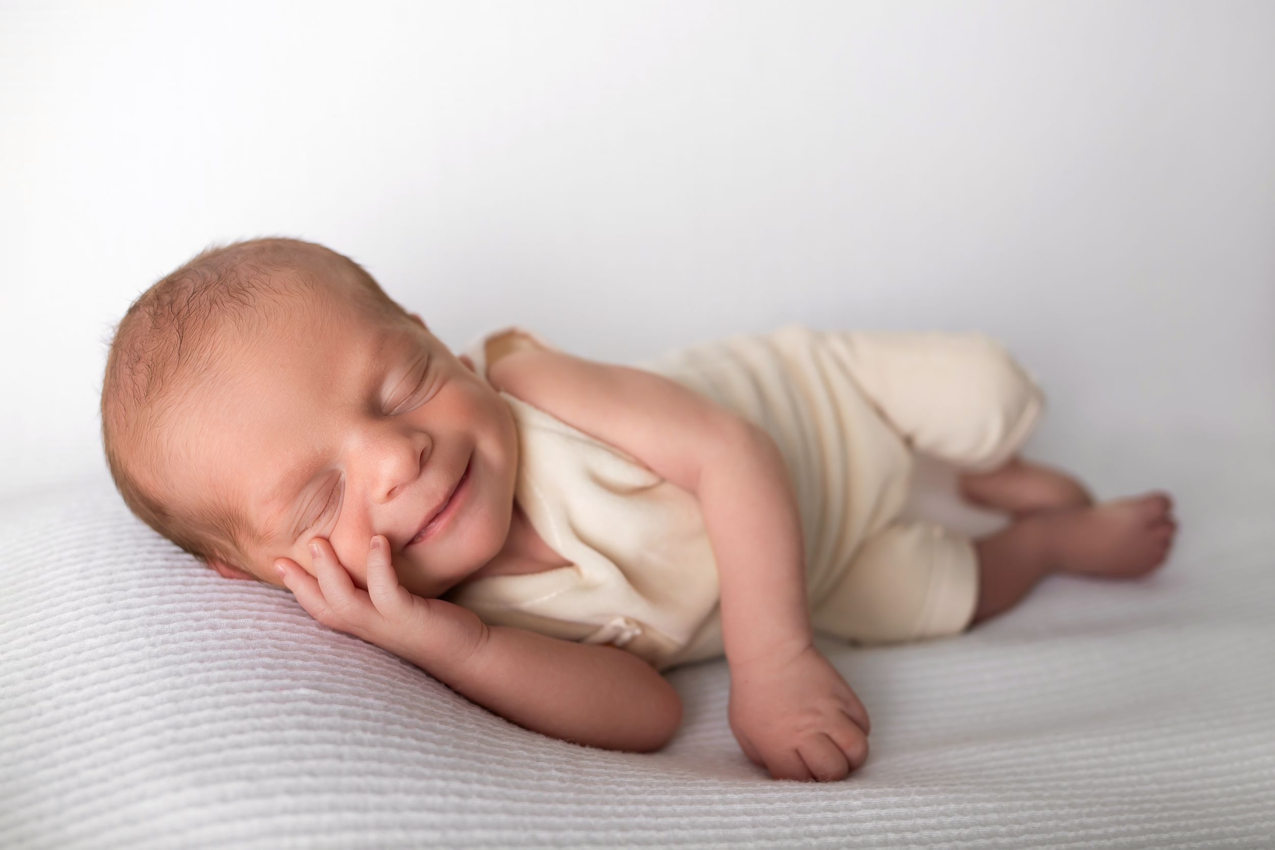 Sleeping baby in cream outfit lying on side on white backdrop.