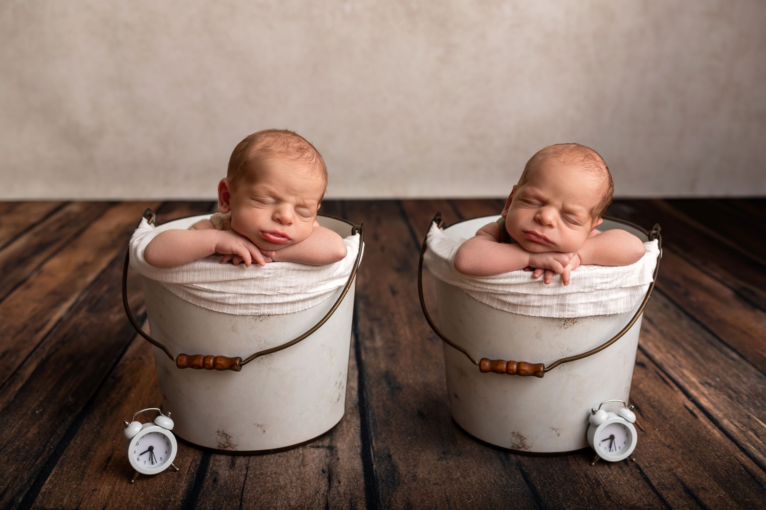 Two sleeping newborns posed in matching white buckets on wooden floor.