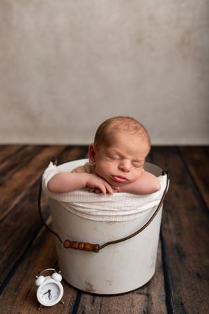 Peaceful newborn sleeping in vintage white bucket with small alarm clock beside.