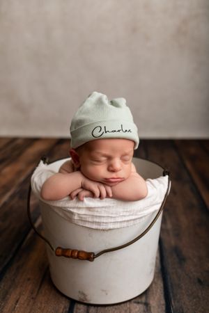 Newborn in monogrammed beanie sleeping atop white bucket on wooden floor.