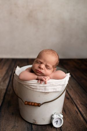Newborn peacefully sleeping on white cushion in rustic bucket with alarm clock prop.