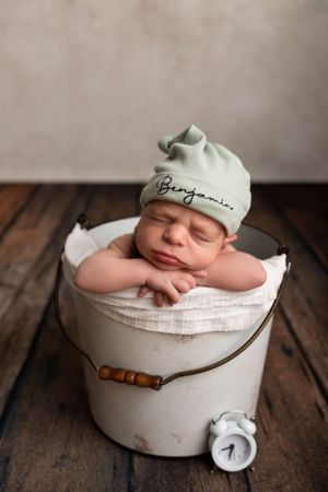 Sleeping newborn in a mint green hat rests in a vintage white bucket on wooden floor.