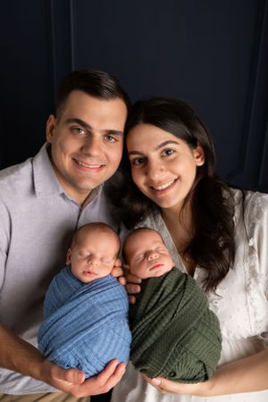 Family portrait with parents holding twin babies wrapped in blue and green swaddles against dark background.