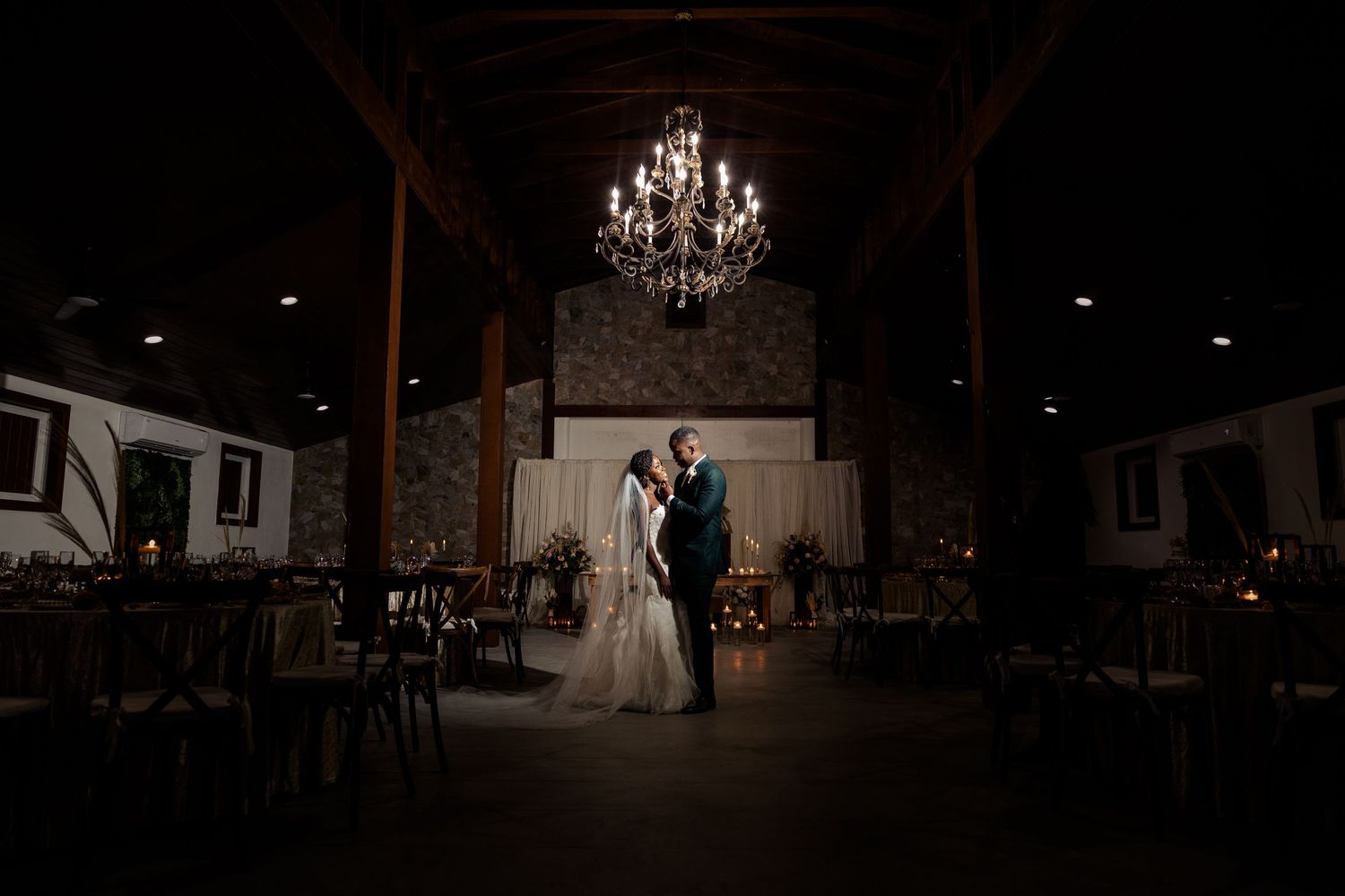 A couple stands in a dark reception hall illuminated by an elegant chandelier during their wedding celebration.