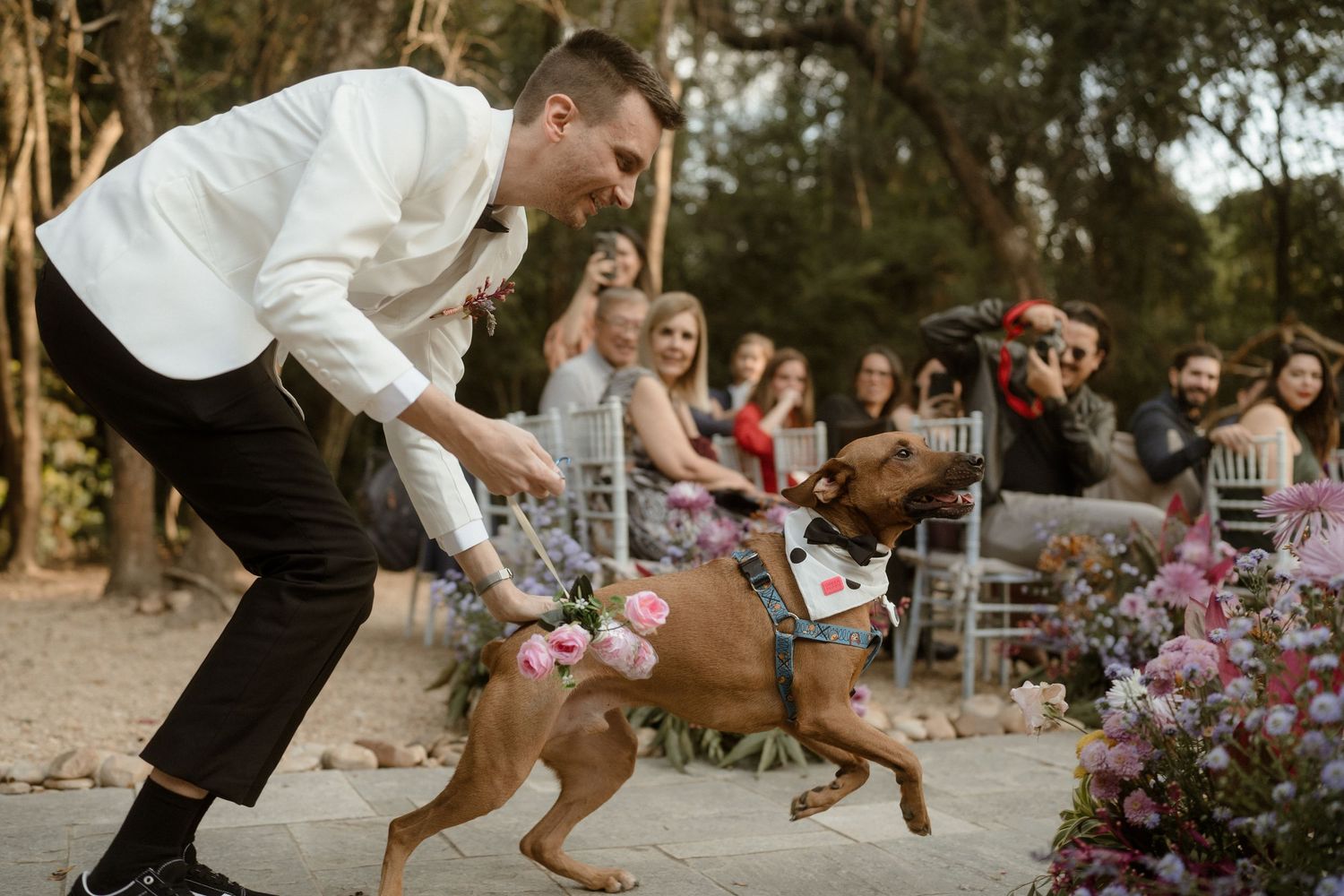 A playful moment with a dog during an outdoor wedding celebration surrounded by floral decorations.