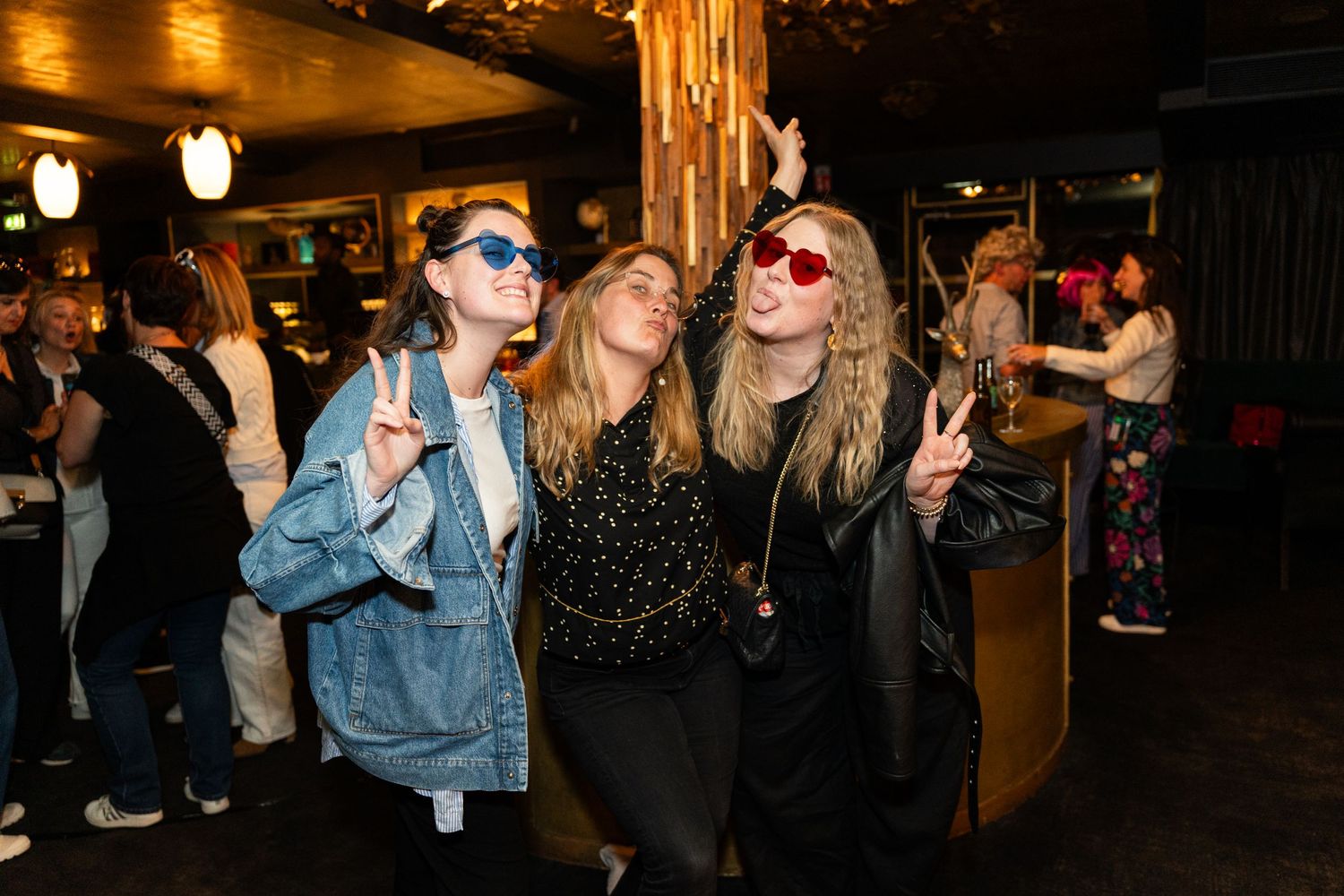 Party goers in sunglasses and trendy outfits dance and pose together at a dimly lit nightclub event.