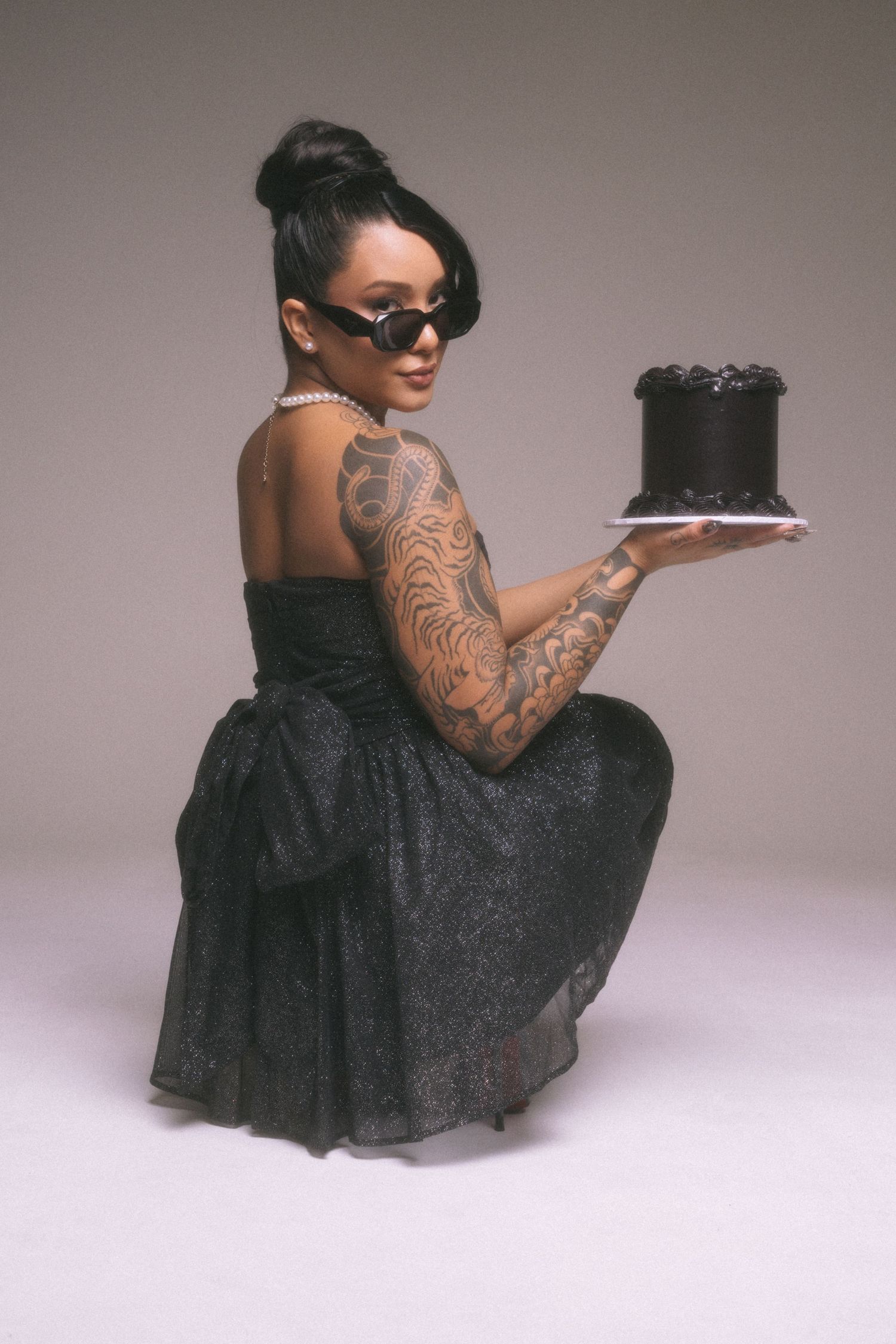 A person in a black dress and sunglasses poses with a dark cake, showing off intricate arm tattoos in a studio setting.