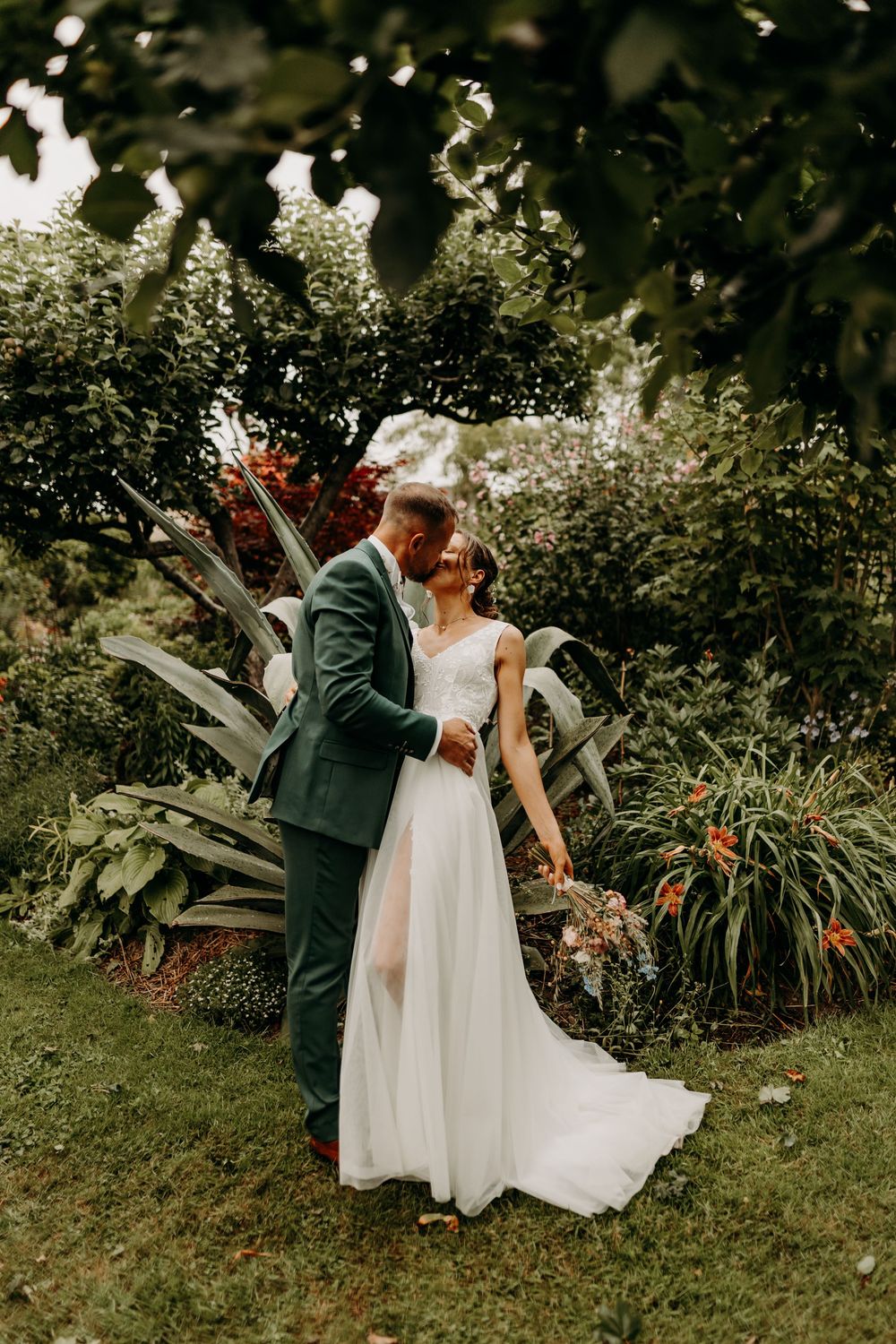 A couple in wedding attire shares an intimate moment in a lush garden setting with blooming flowers and greenery.