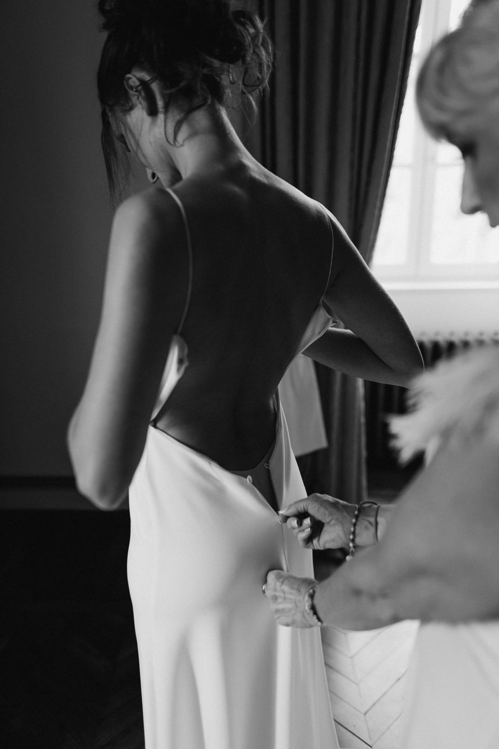Black and white photo of someone getting dressed in an elegant white gown with a bare back being zipped up.