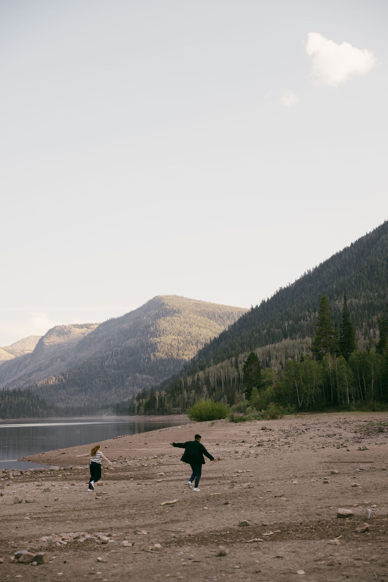 Two people running freely on a beach shore with mountains and lake in the background.