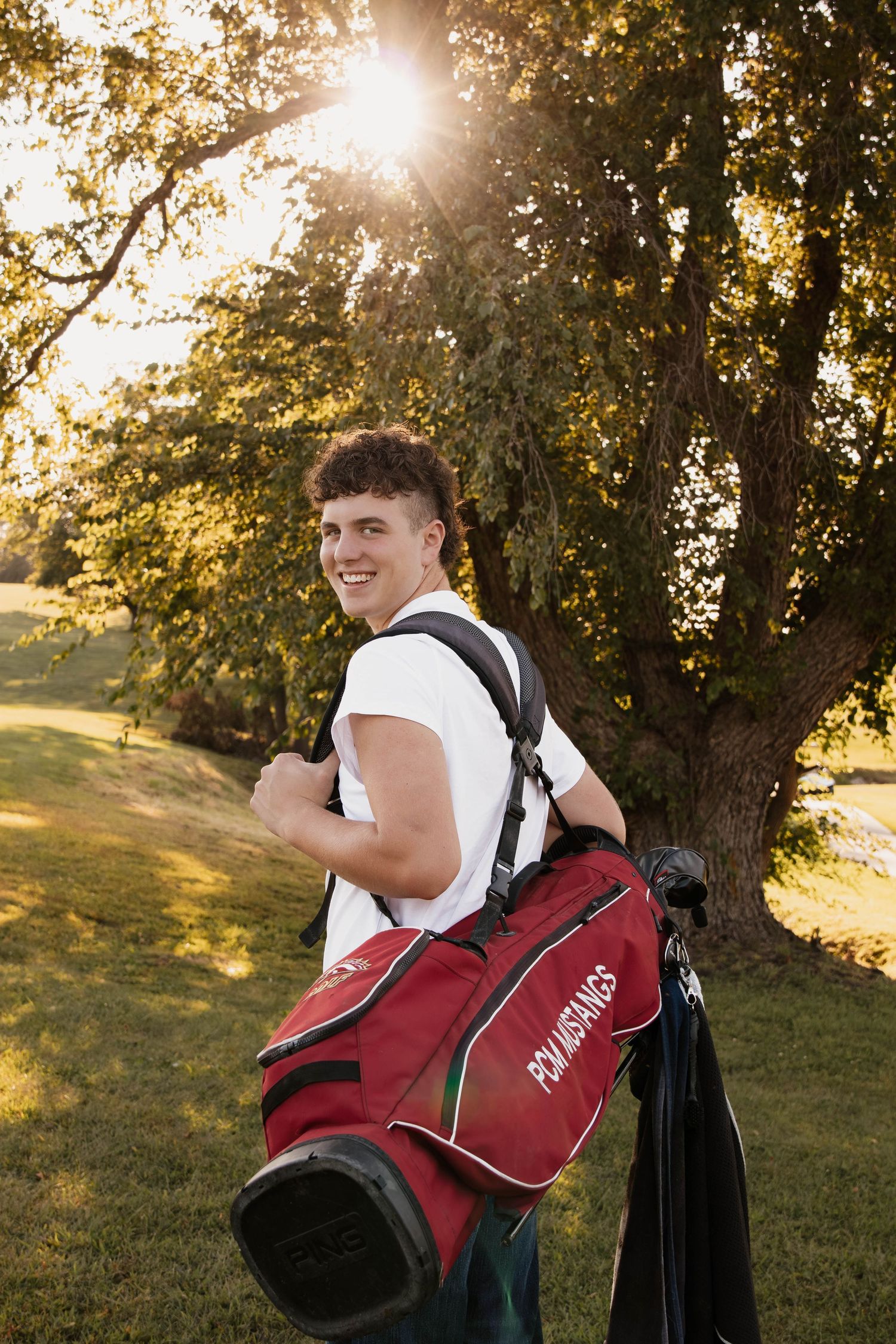 Golfer carrying red golf bag smiles while walking on course at sunset.