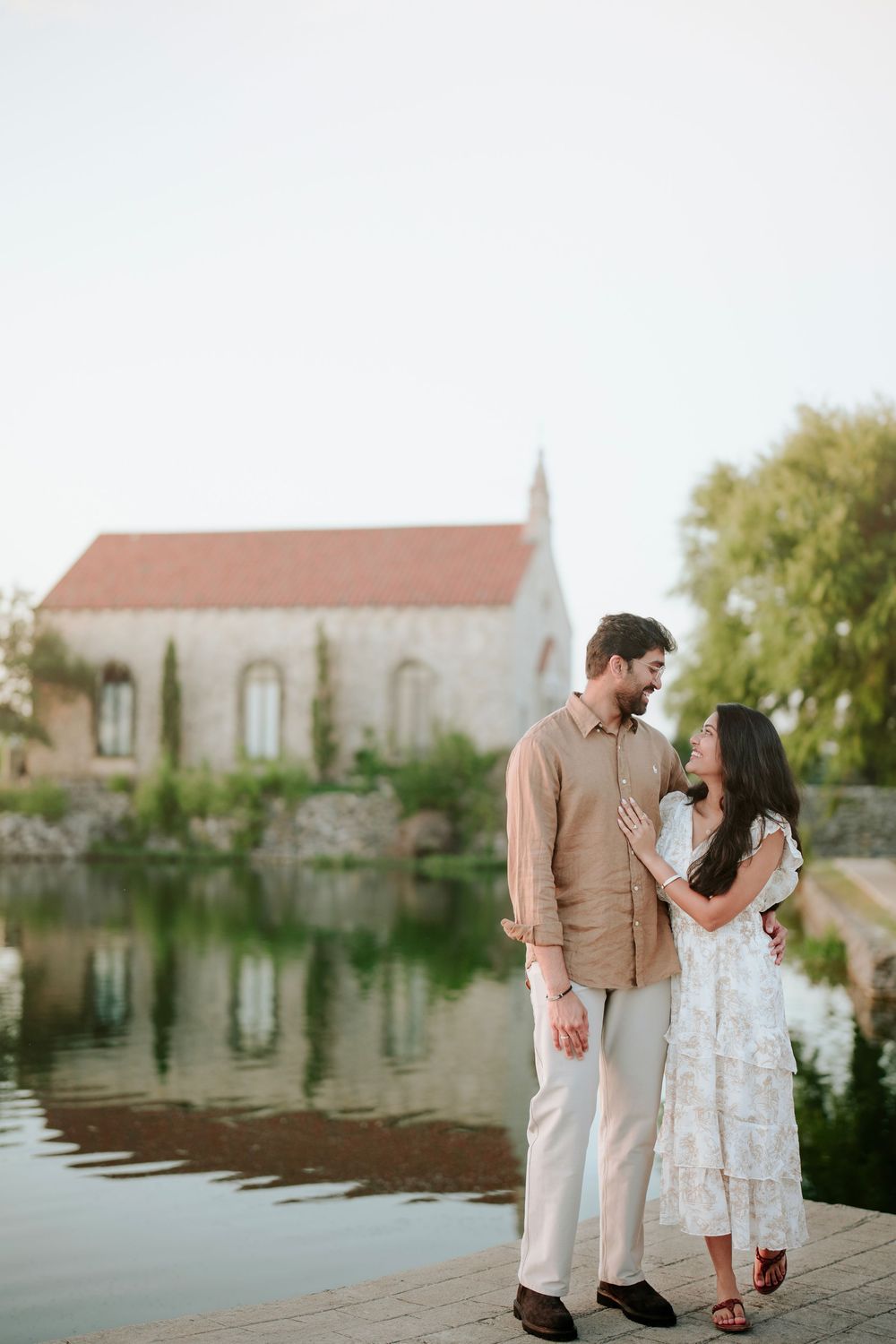 Couple in elegant attire standing by a reflective pond with a historic stone chapel and trees in background.