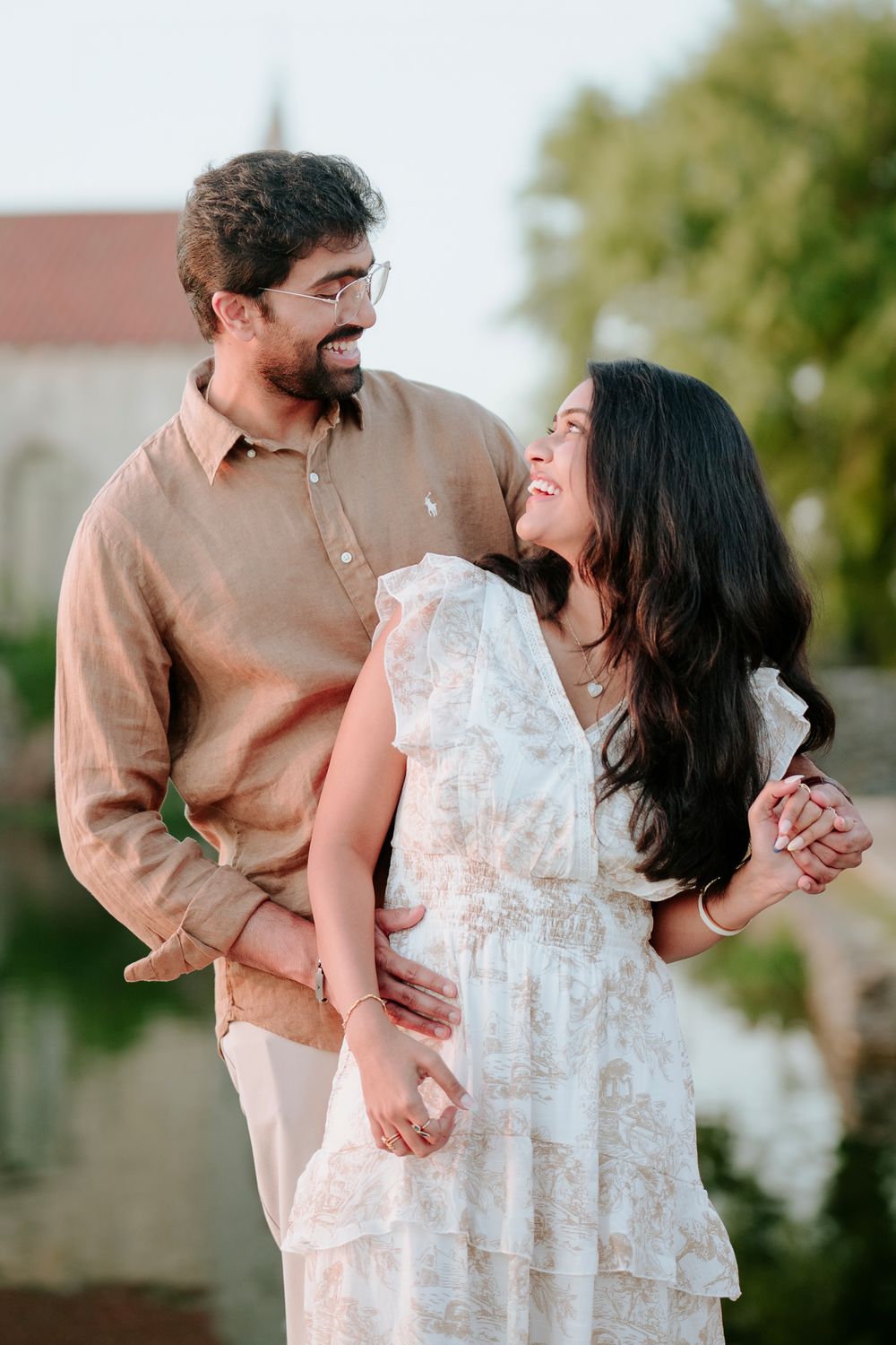 A smiling couple shares a joyful moment outdoors, man in tan shirt embracing woman in white floral dress.