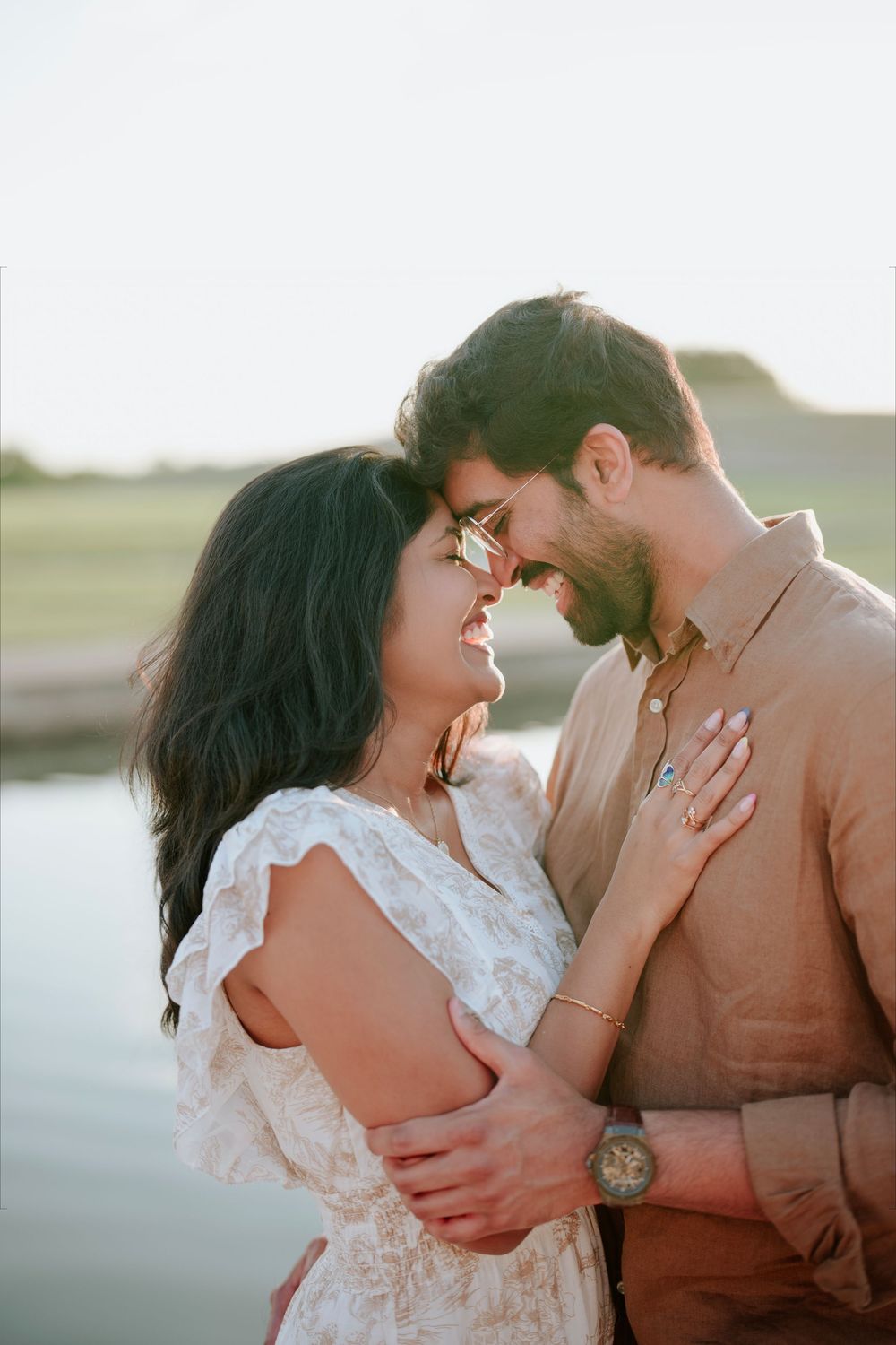 Romantic couple embracing and smiling at each other near a serene waterfront at golden hour.