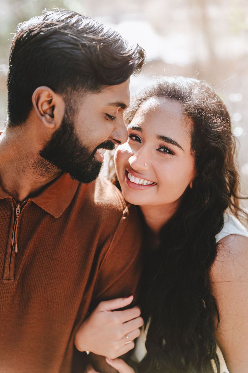 A smiling young couple sharing a romantic moment outdoors, the woman wearing a nose ring and the man in a brown polo shirt.