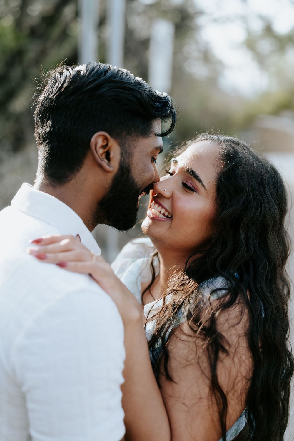 A romantic couple shares an intimate moment, almost kissing, as the smiling woman embraces the bearded man outdoors.