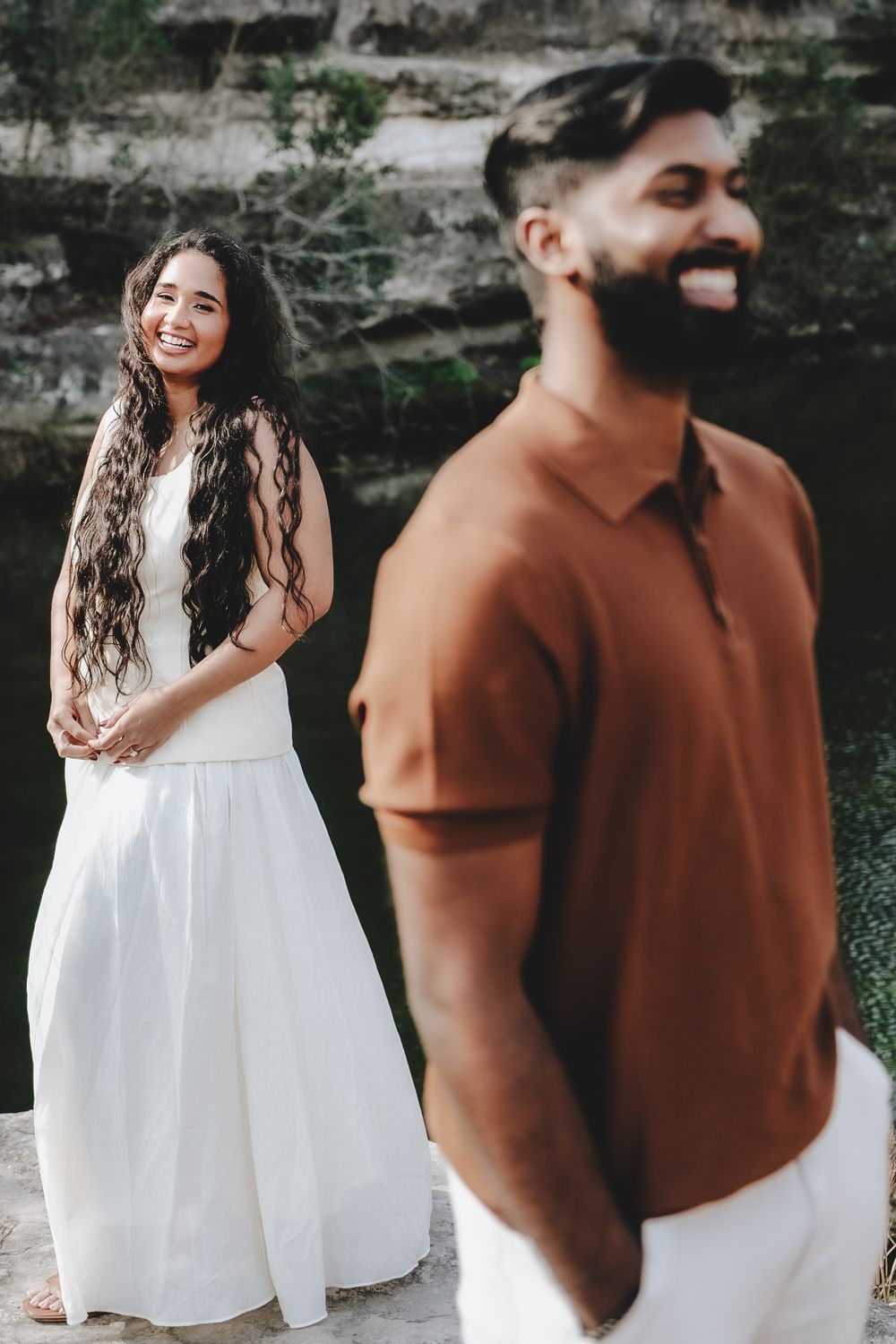A smiling woman in a white dress stands behind a bearded man in a brown polo shirt outdoors near rocky cliffs.