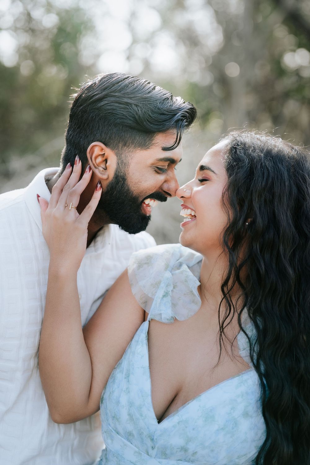 A smiling couple shares a romantic moment outdoors, with the woman tenderly holding the mans face as they lean in close.