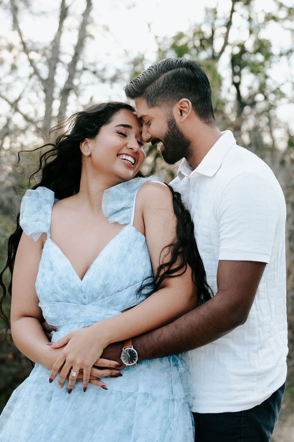 A smiling couple embraces outdoors, woman in light blue dress, man in white shirt, touching foreheads romantically.