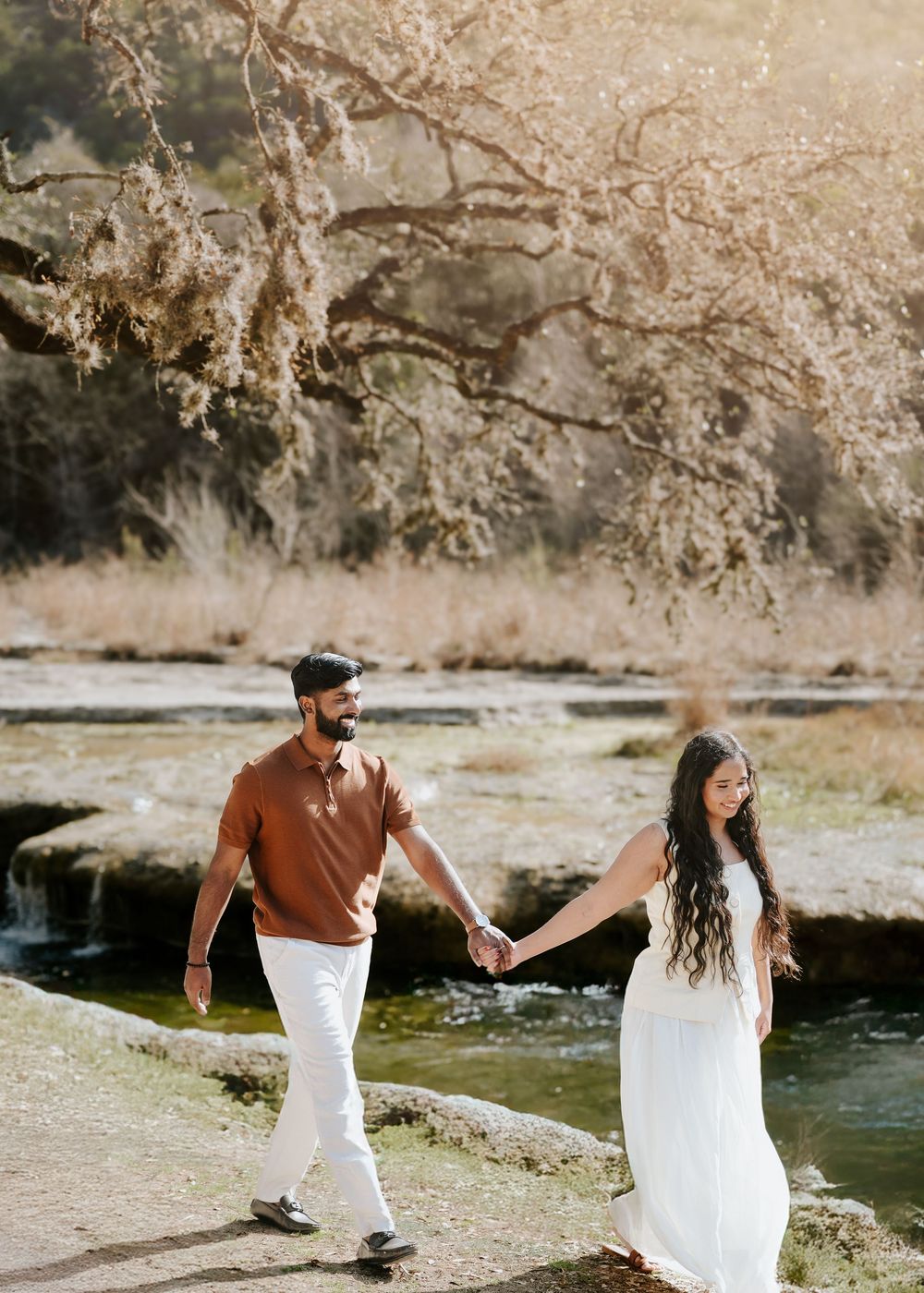 Couple holding hands walking near a sunlit creek, woman in white dress and man in brown polo shirt.