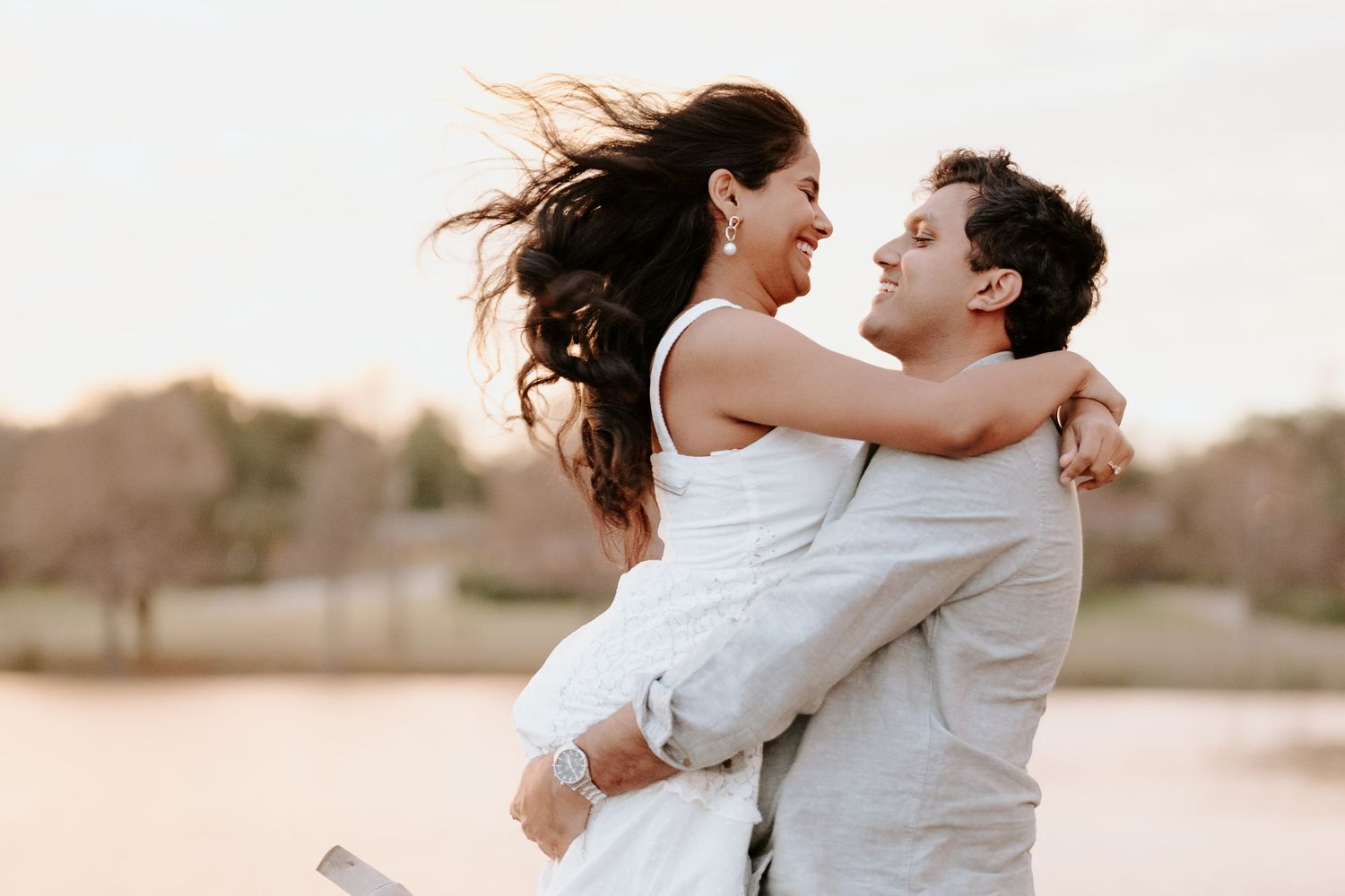A couple in wedding attire embrace and laugh together during sunset, with the bride's hair flowing in the wind.