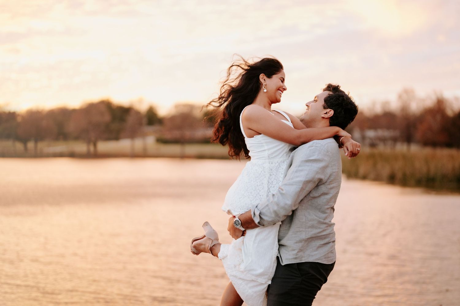 A couple embraces joyfully in a field at sunset as her white dress flows in the gentle breeze.