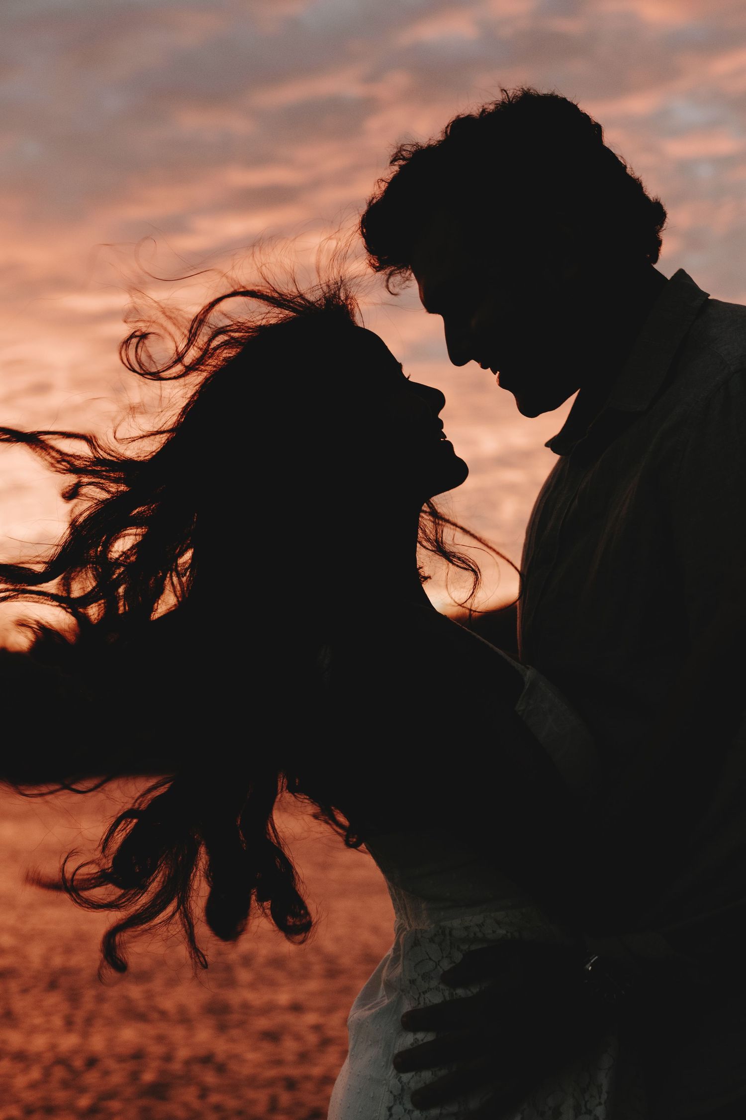Silhouette of a romantic couple against a dramatic orange sunset sky with flowing hair in the wind.