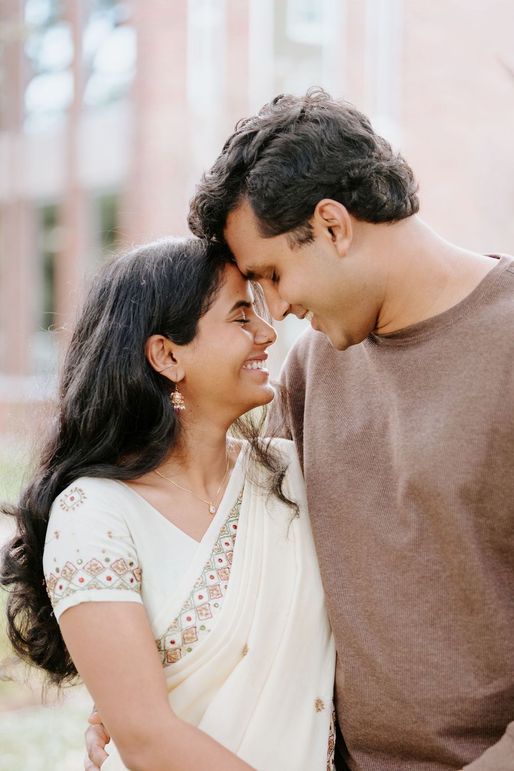A smiling South Asian couple in traditional and casual attire touch foreheads tenderly outdoors.