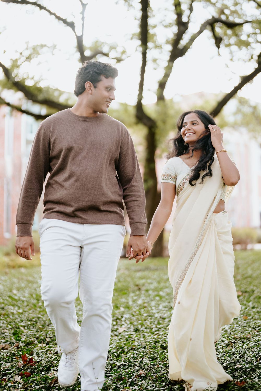 A smiling couple walks hand-in-hand outdoors, man in brown sweater and woman in elegant cream saree, with trees in background.