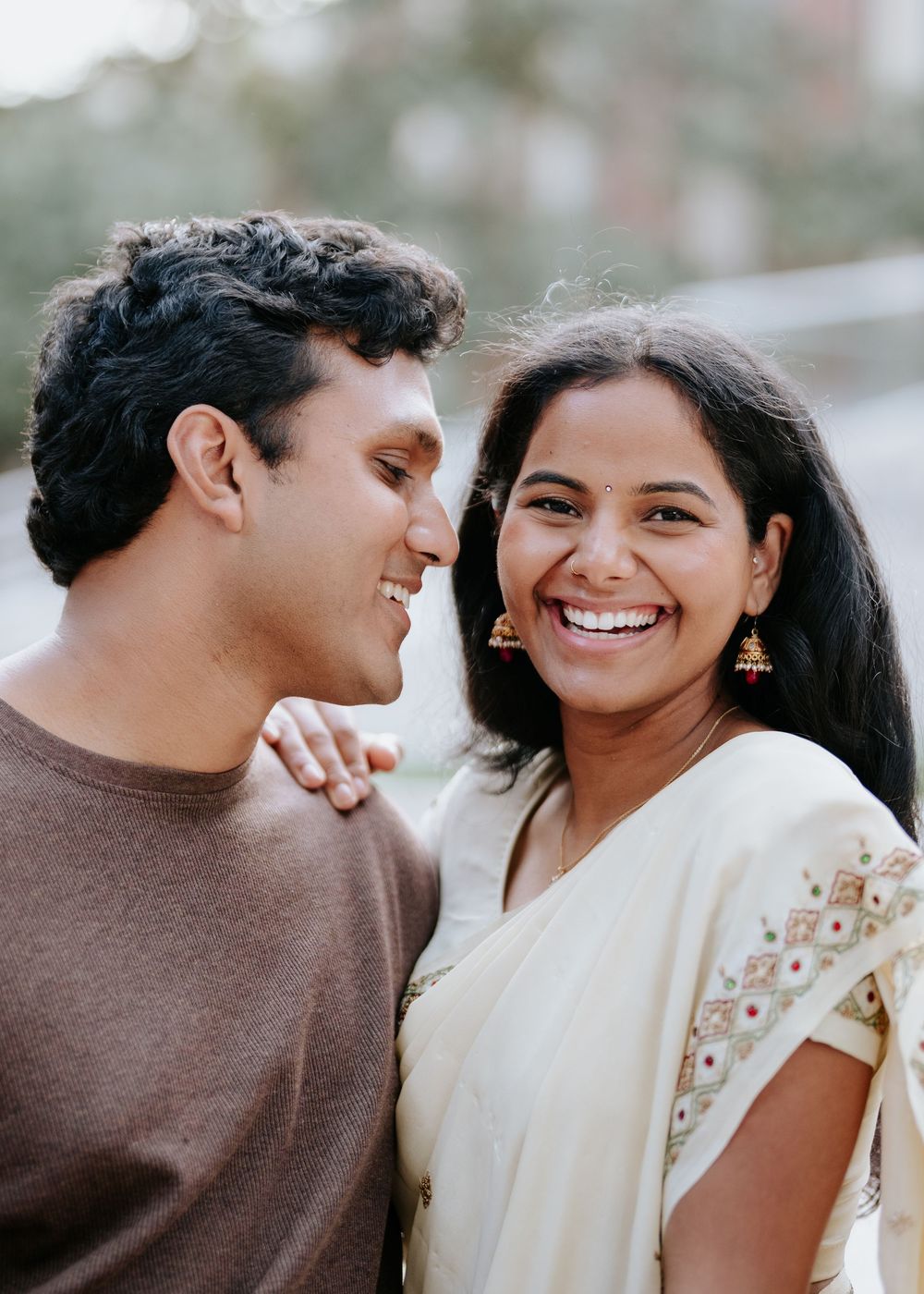 A smiling South Asian couple in casual and traditional attire share a joyful moment together outdoors.