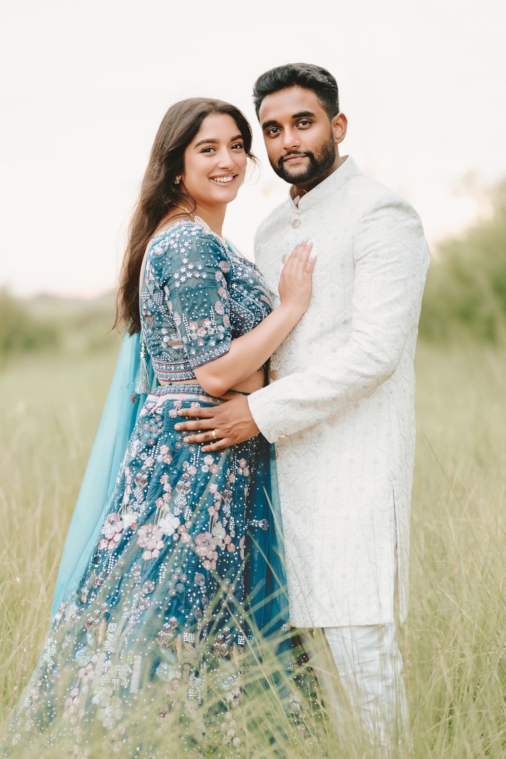 Couple in traditional Indian attire poses in a grassy field, woman in blue lehenga and man in white sherwani.