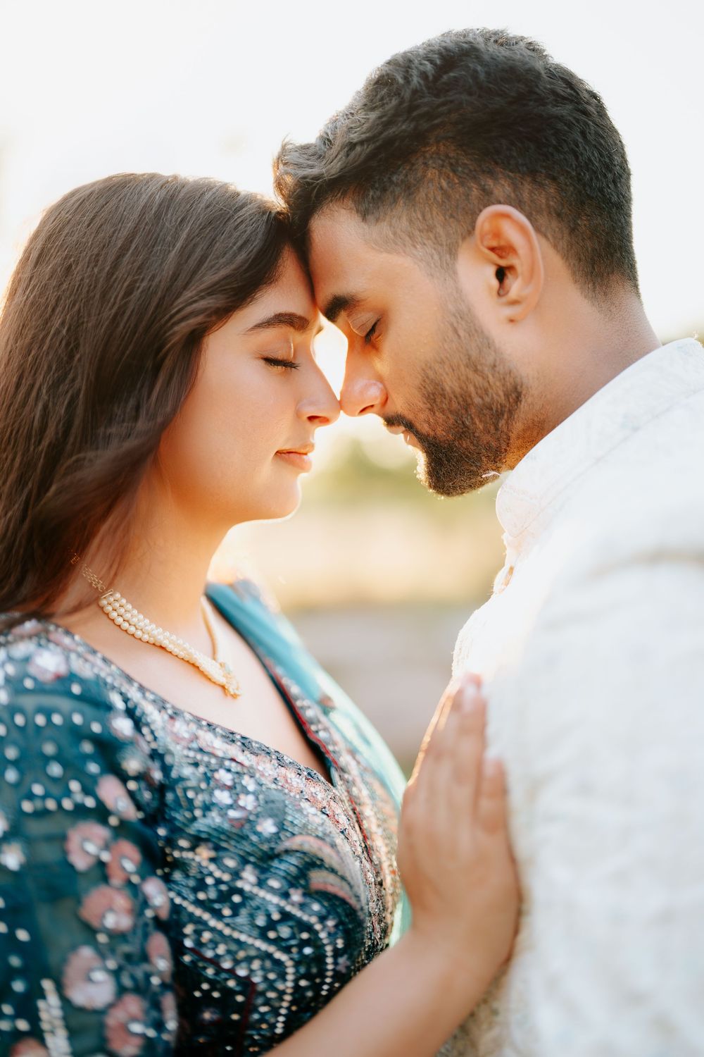 A romantic South Asian couple in traditional attire touch foreheads tenderly in a soft golden light outdoor setting.