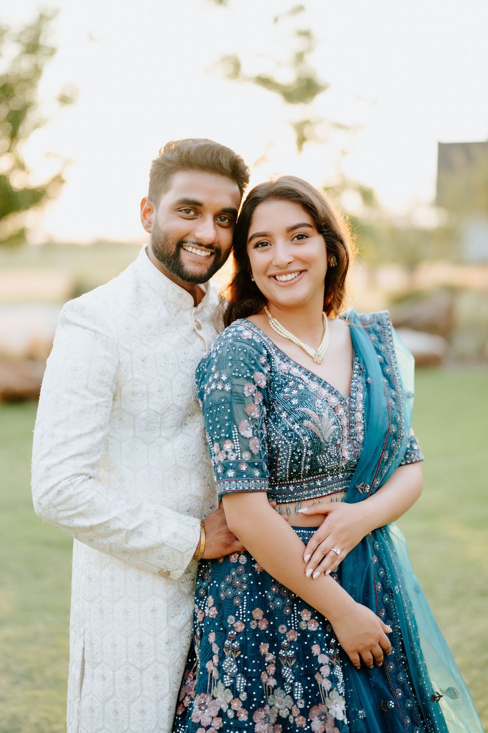 A smiling couple in traditional Indian attire, man in white sherwani and woman in blue floral lehenga, posing outdoors at sunset.