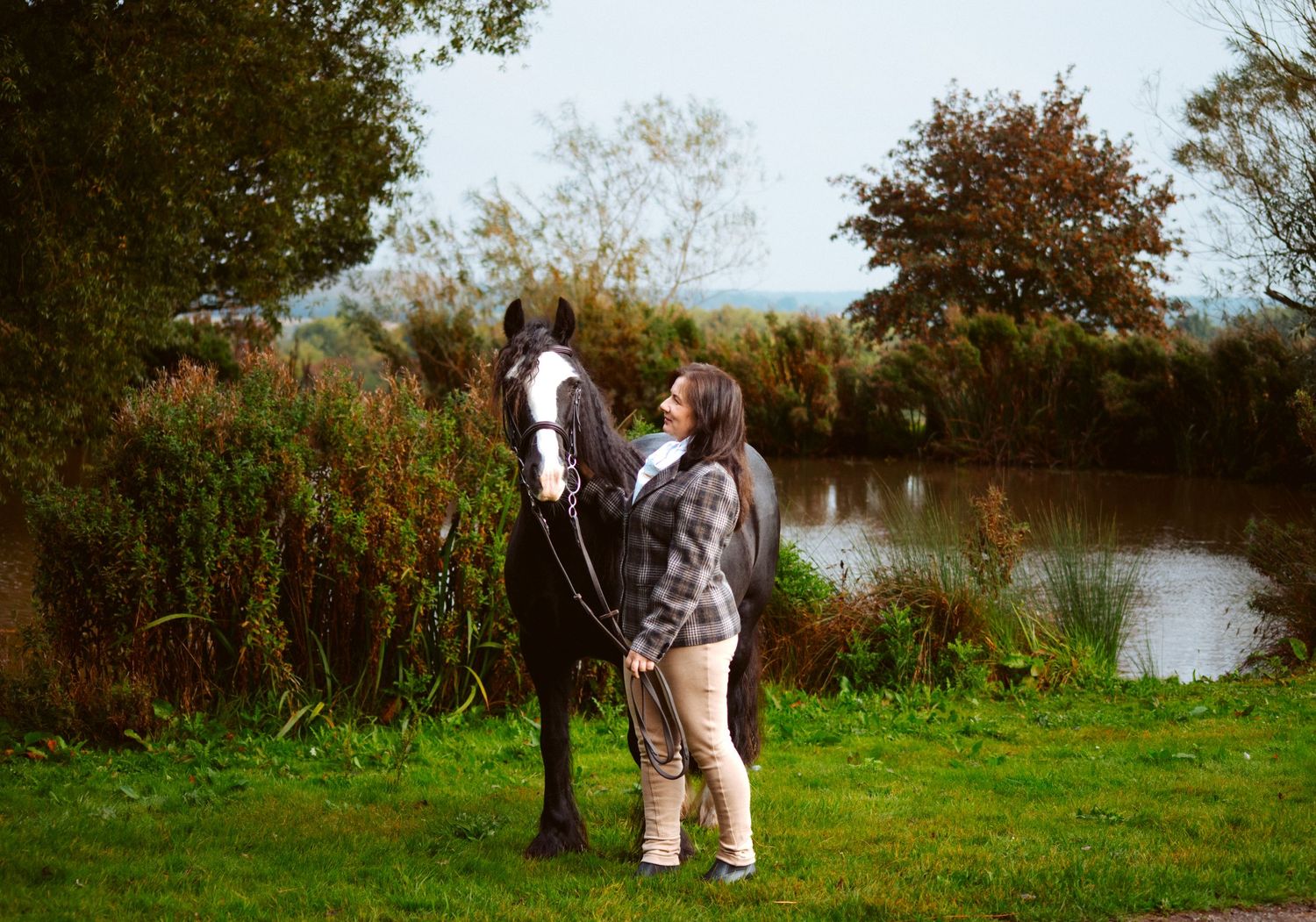 Two horses and their handler beside a tranquil pond on a fall day.