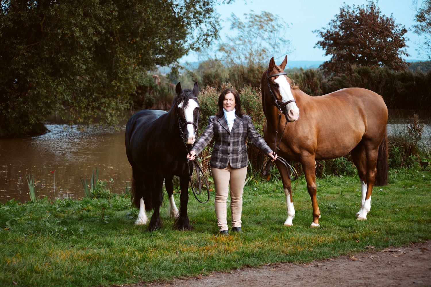 Two horses flanking their handler in a plaid jacket by a pond.