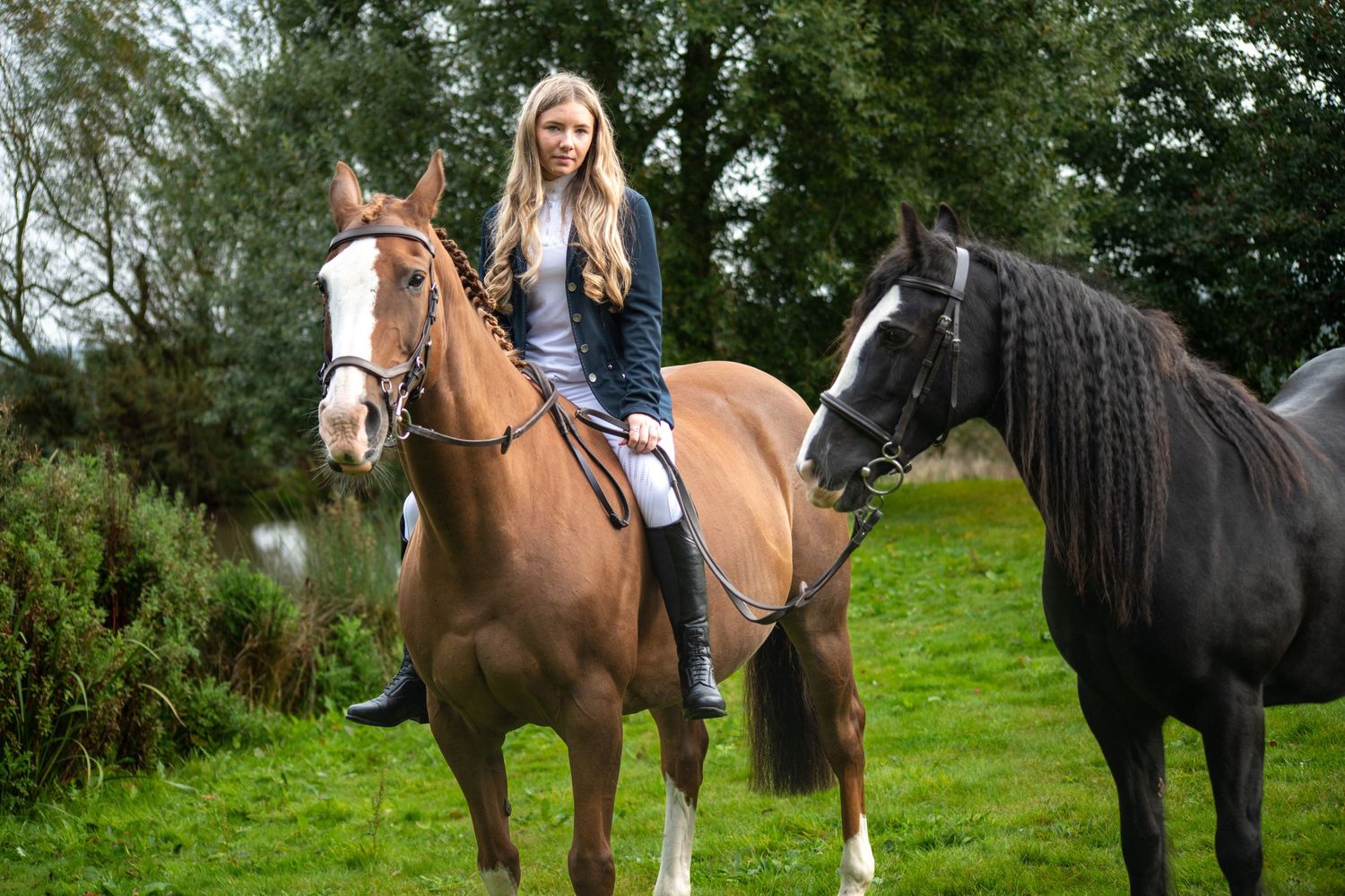 A rider on a chestnut horse in a grassy field with a black horse standing nearby.