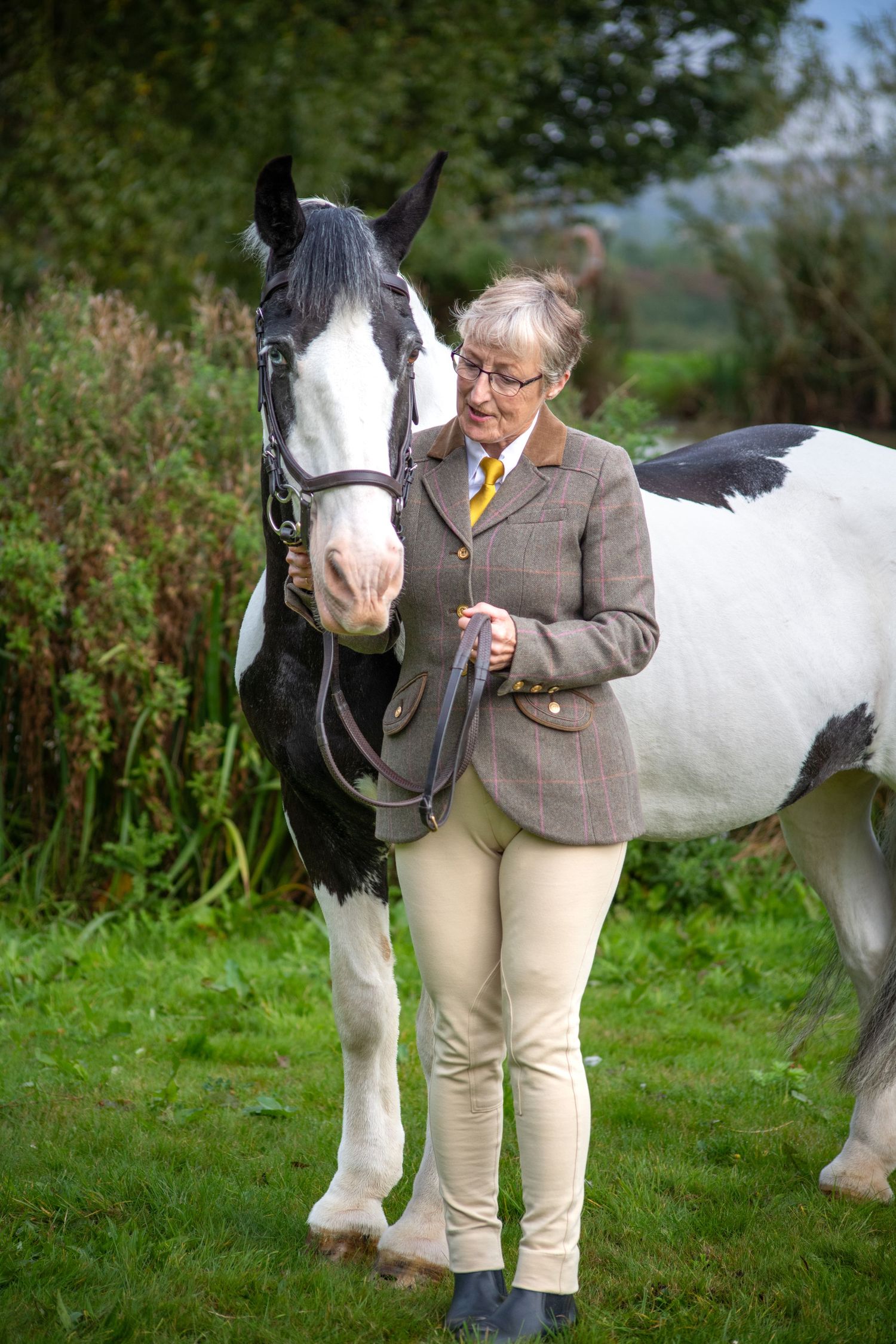 Person in tweed jacket leading black and white horse through green countryside.