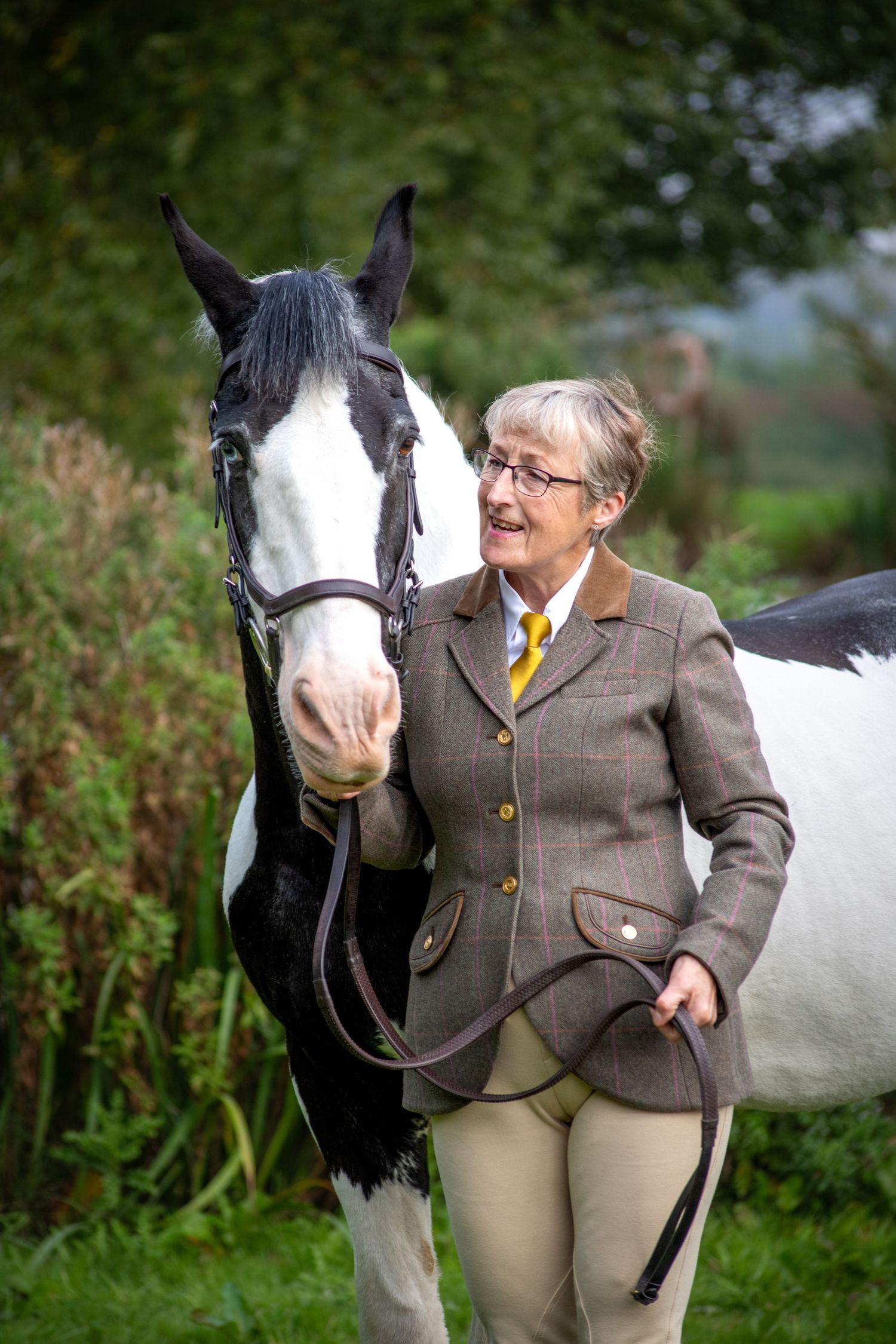 A black and white horse with a blaze stands beside a person in traditional equestrian attire on a grassy field.
