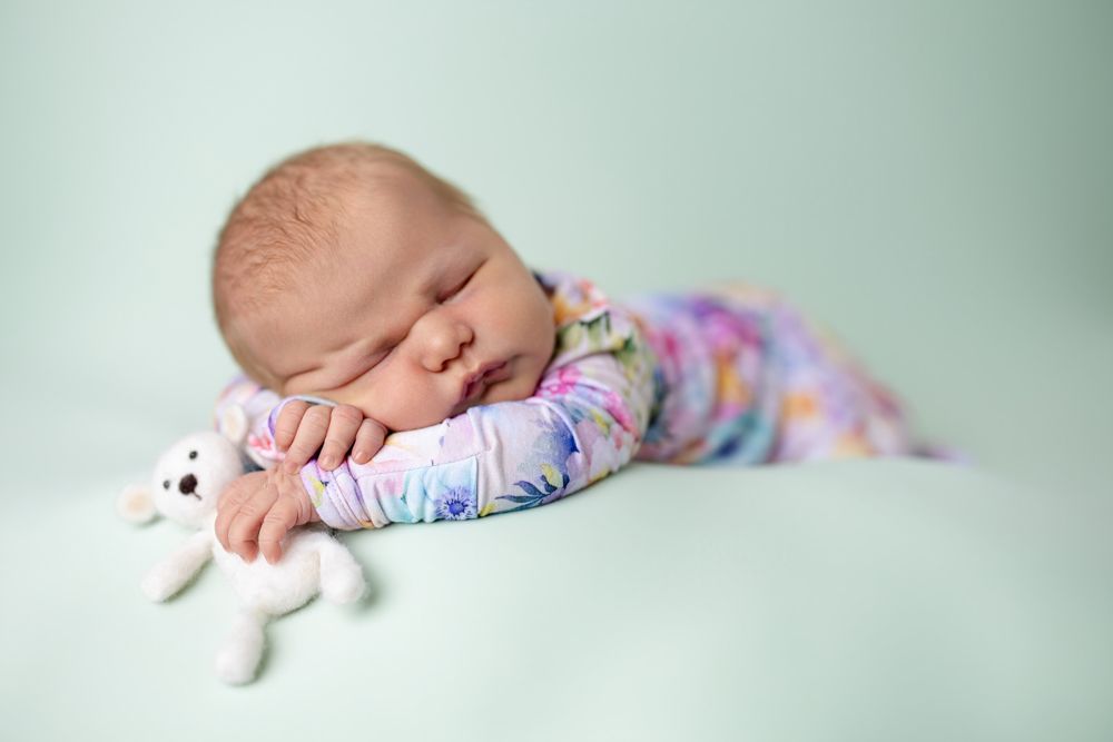 a sleeping newborn in a floral print outfit holding a small stuffed animal on a sage green backdrop