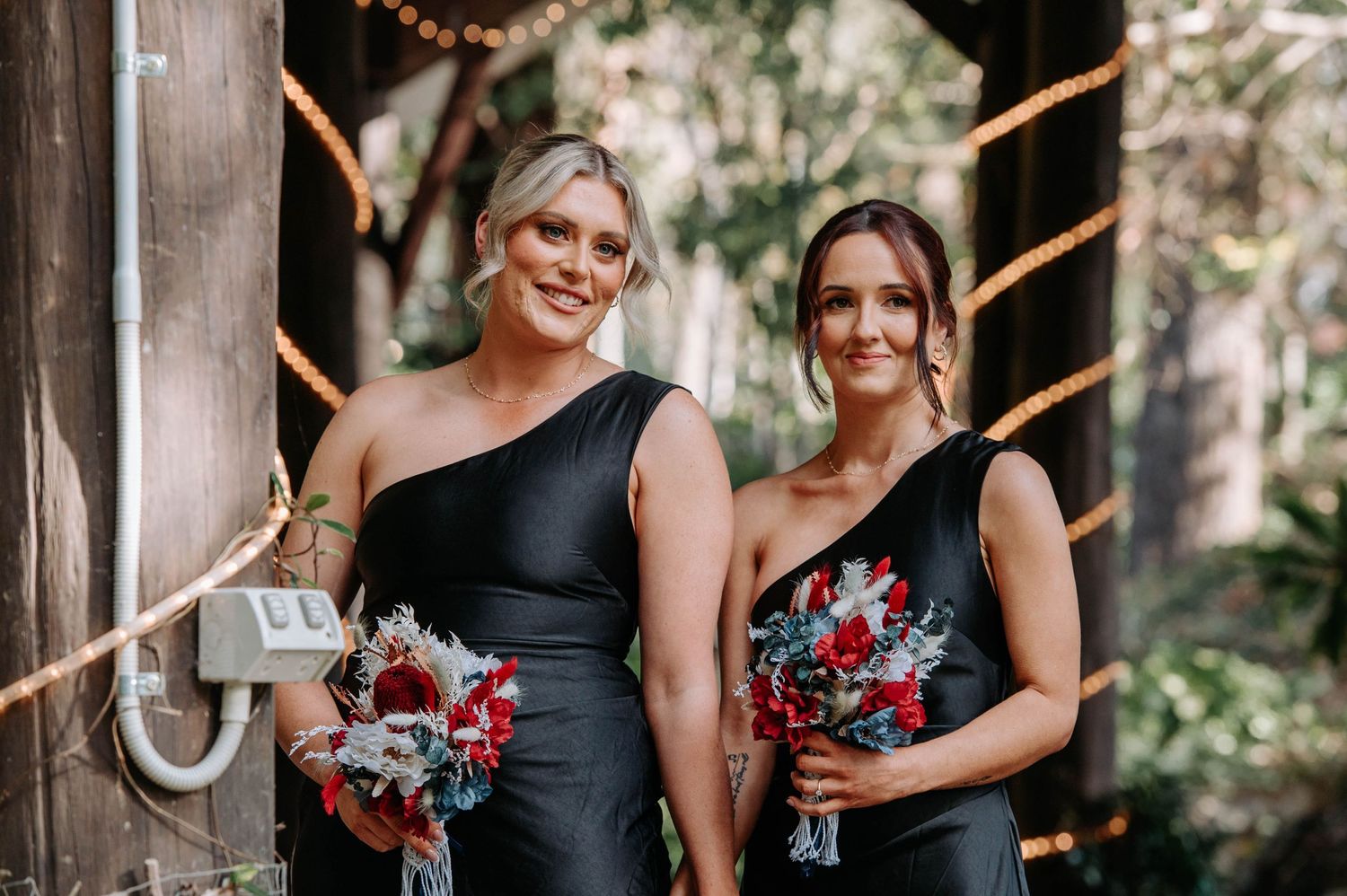 Two bridesmaids in black dresses holding red bouquets posed at outdoor wedding venue.