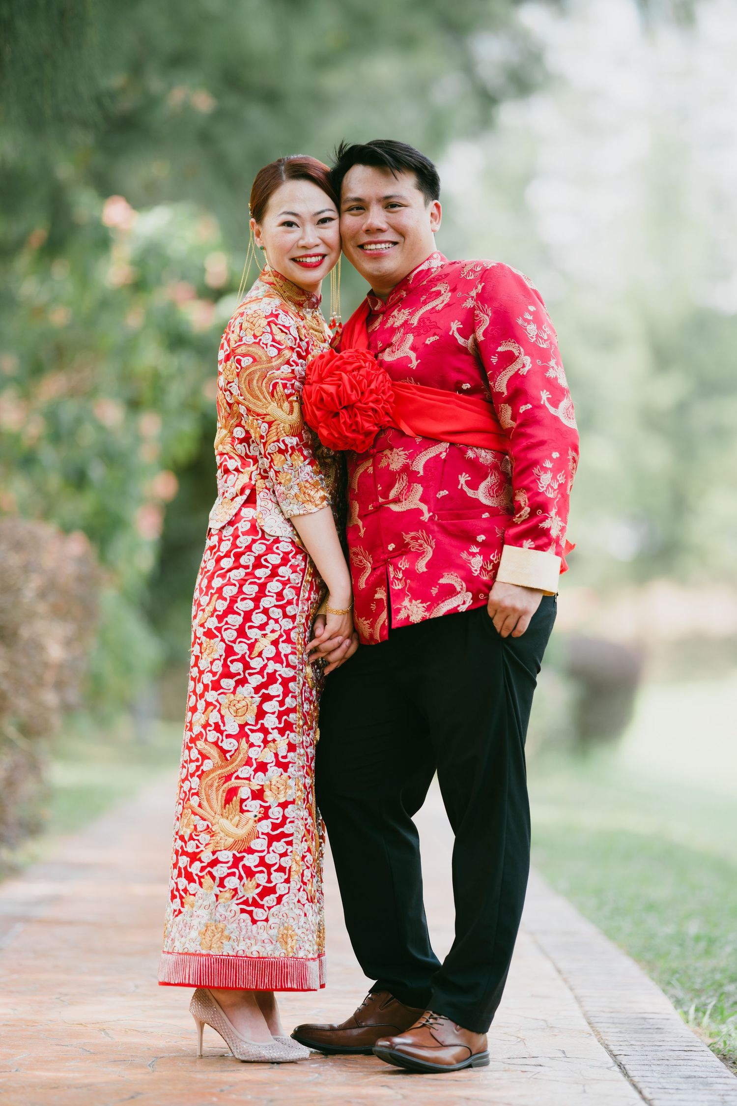 Couple wearing matching red Chinese traditional outfits posing together on an outdoor garden pathway.