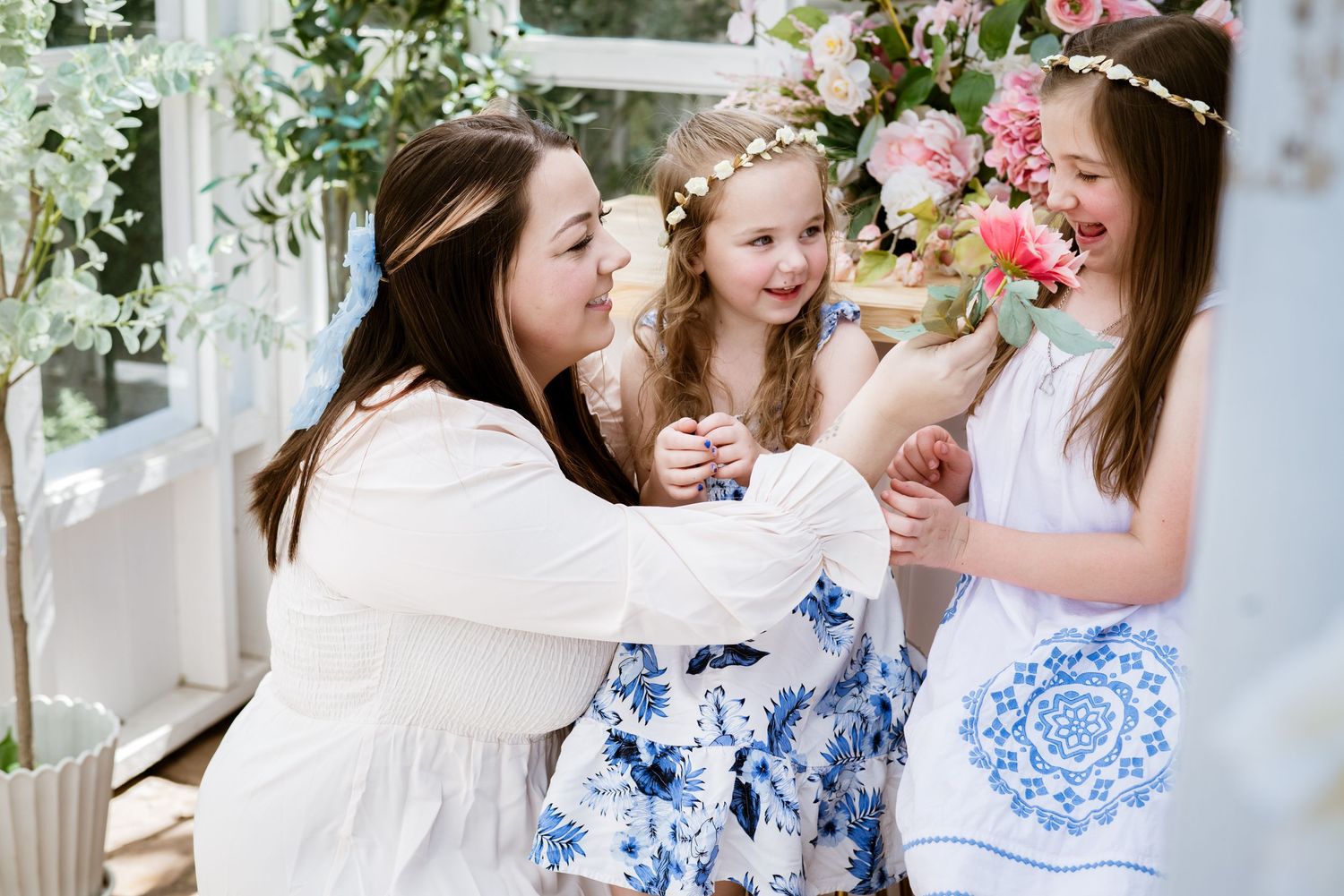 Three women in white dresses share joyful moments together surrounded by pink flowers.