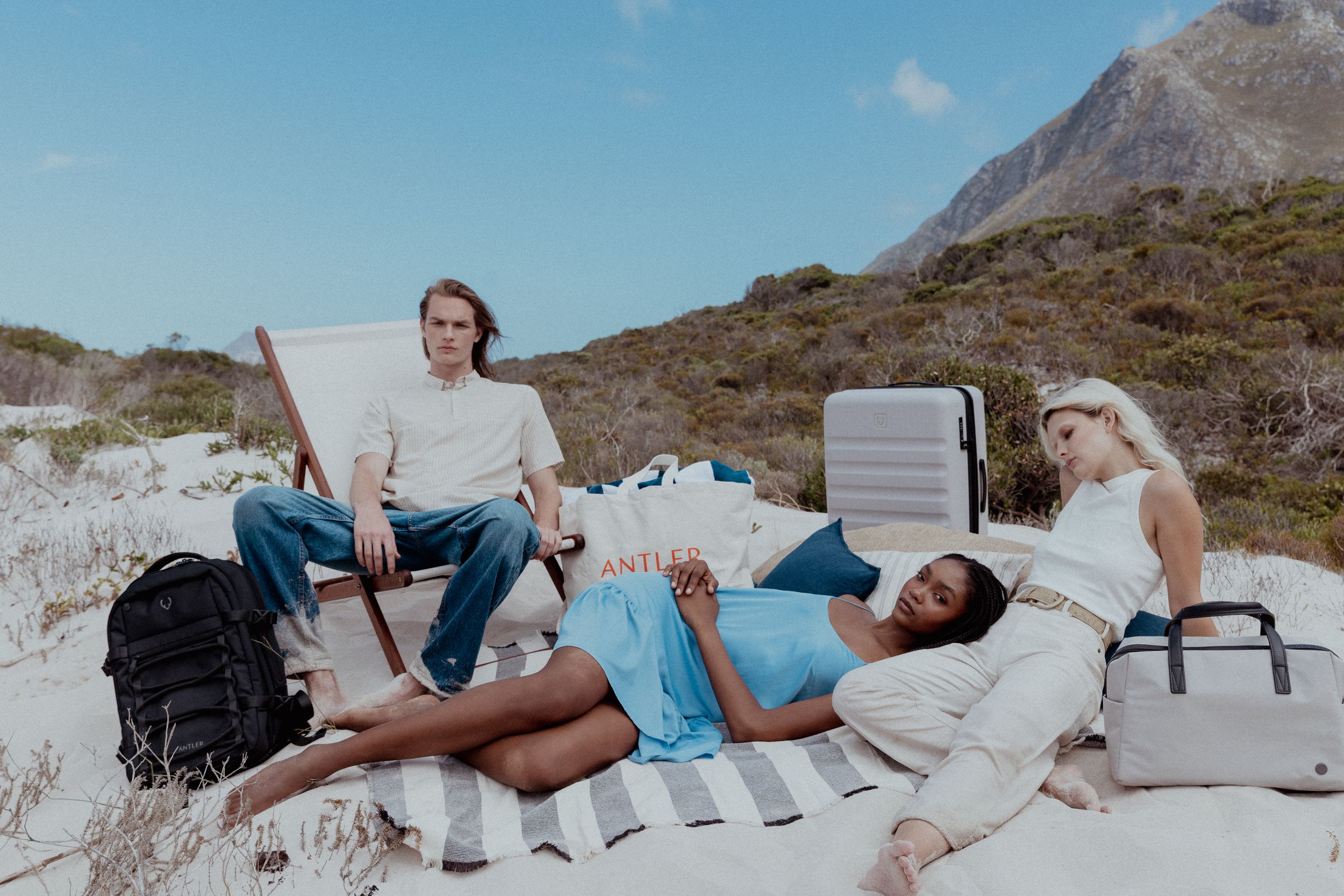 People lounging on beach blankets with luggage amid mountainous coastal landscape.