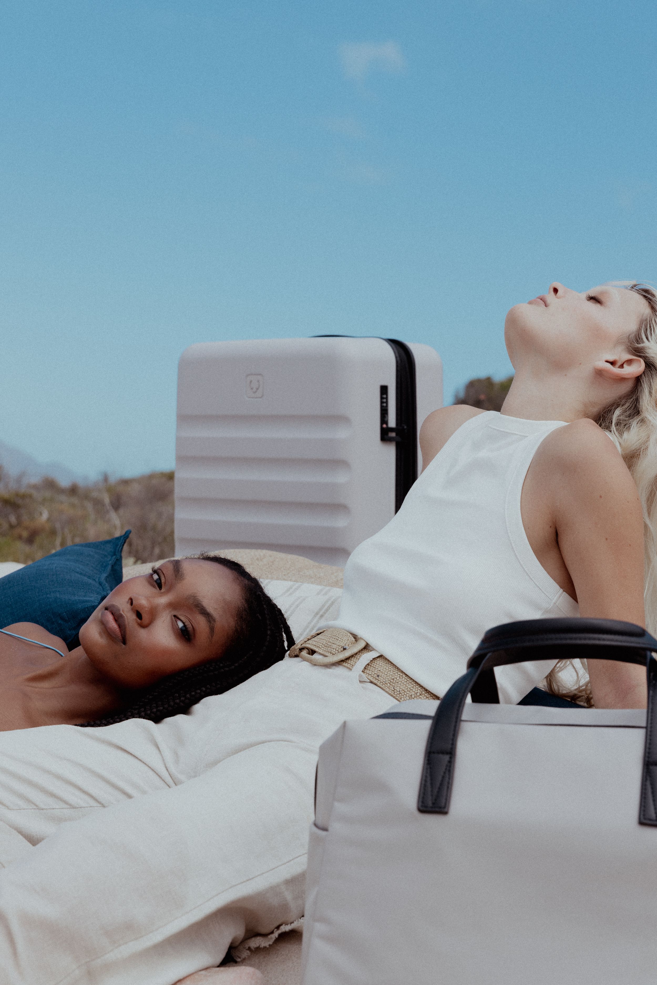Two people relaxing on blanket with white luggage against blue sky backdrop.
