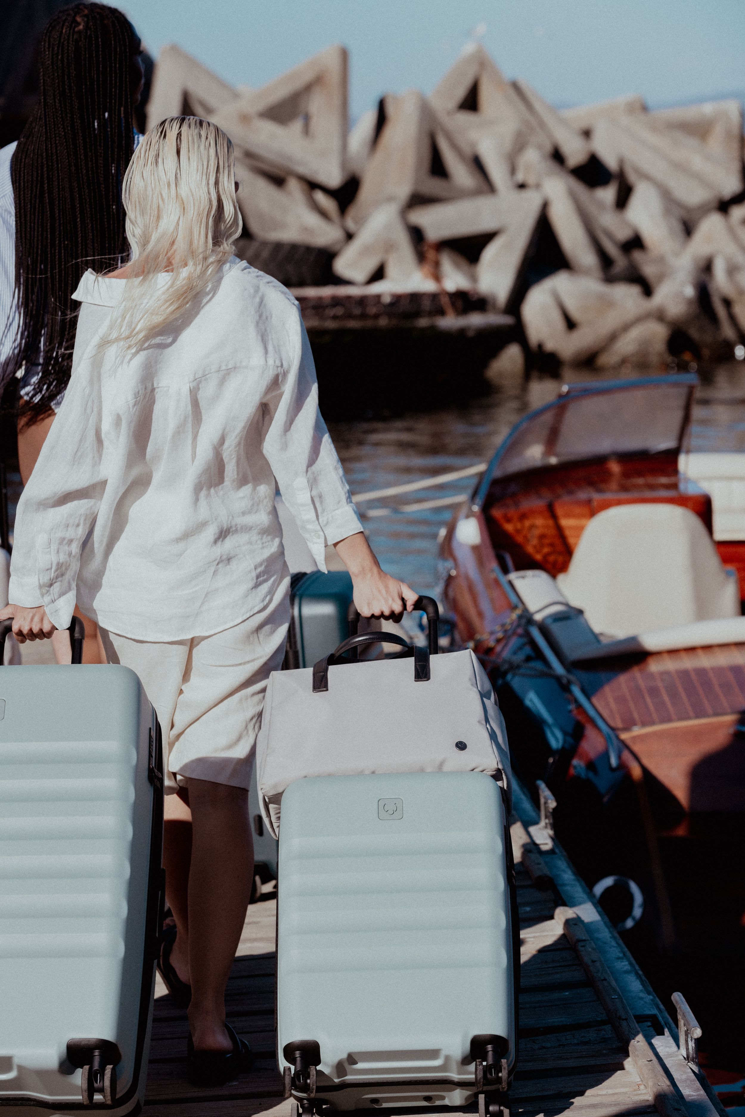 Two people walking with white luggage on dock near geometric stone structures.