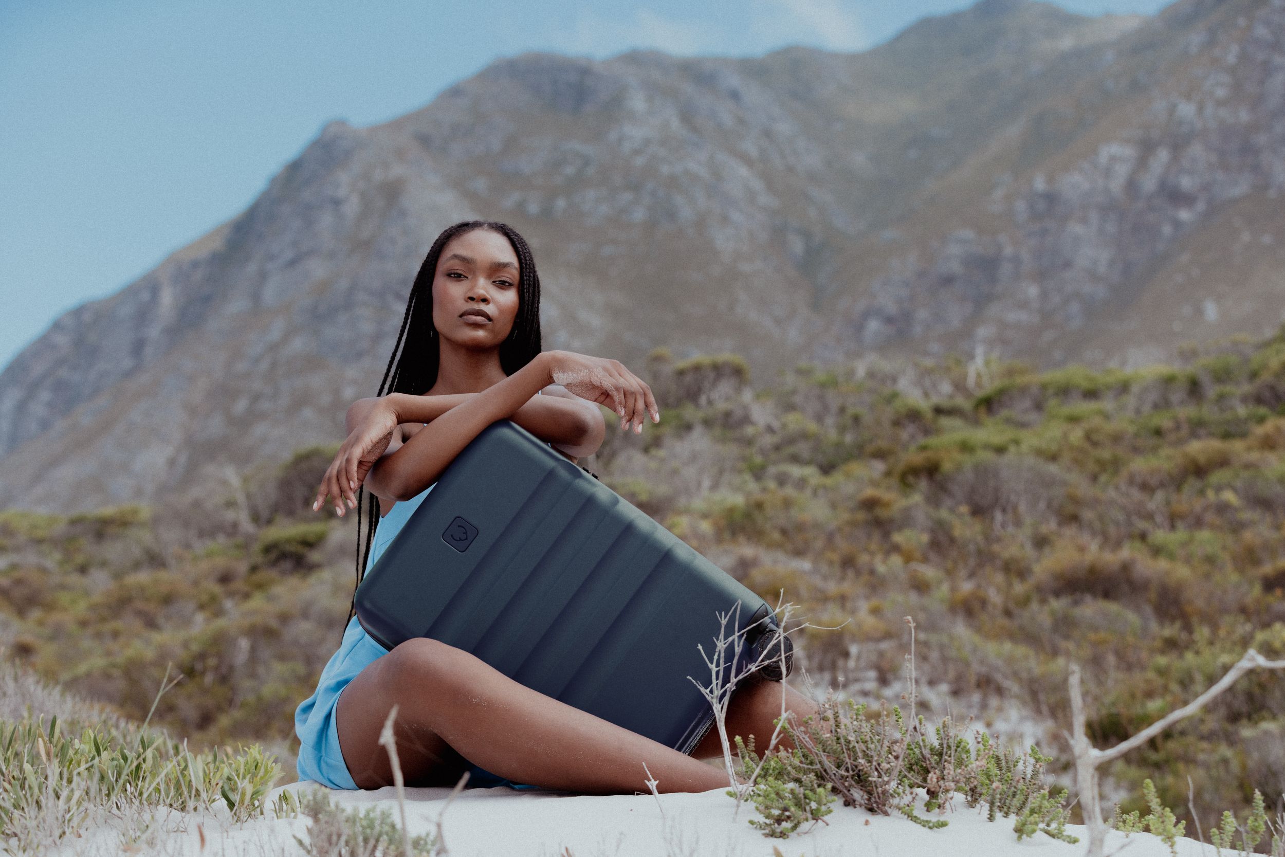 A person poses with luggage against a mountainous backdrop.
