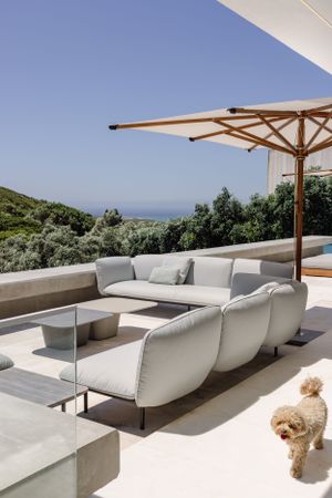 Outdoor terrace with white sectional sofa, wooden umbrella, and coastal views featuring a small dog.