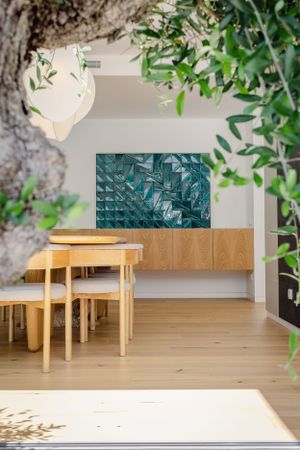 Modern dining area with wooden furniture and turquoise wall art featuring natural light through leafy plants.